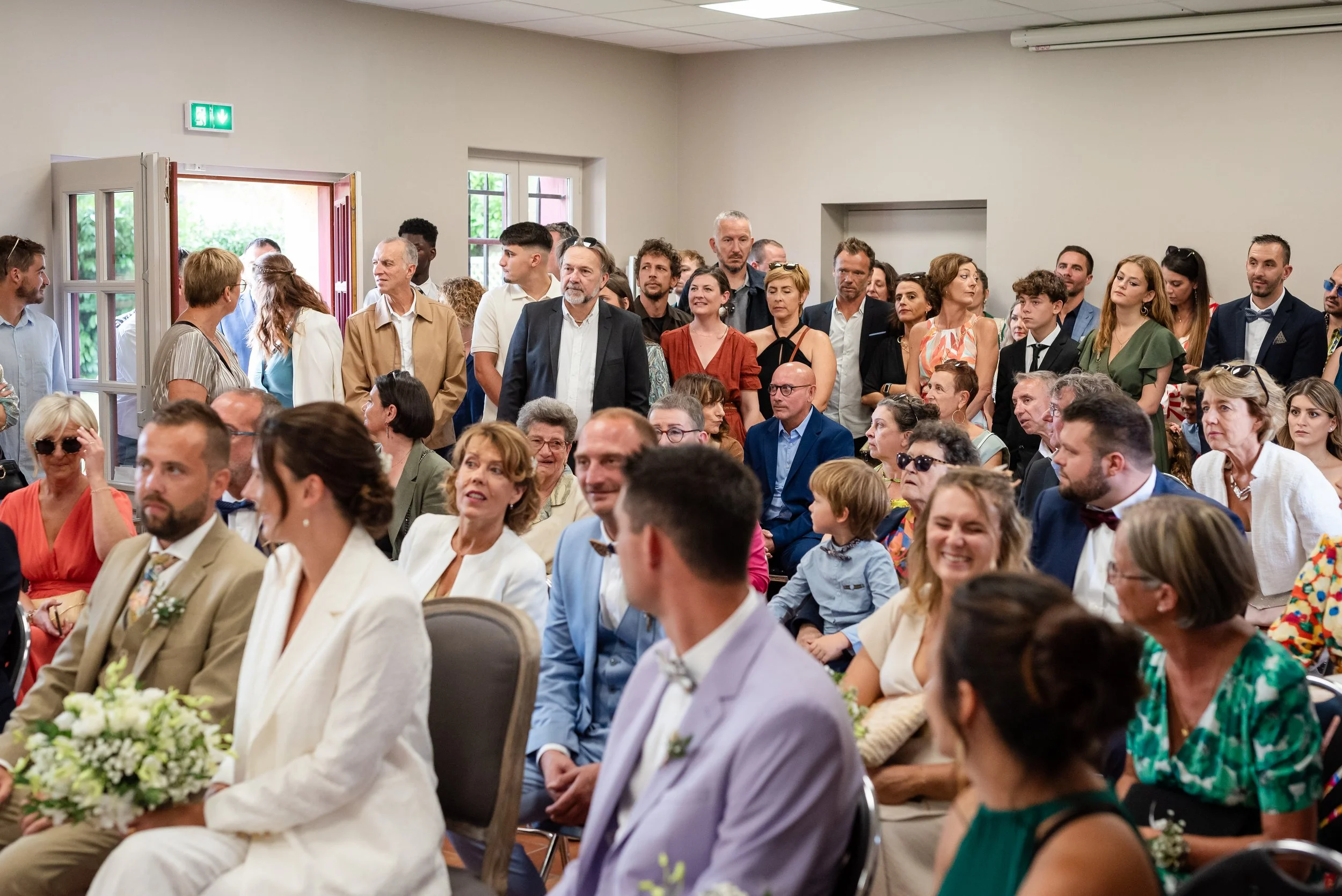 Un groupe de personnes assises et debout dans une salle lors d'un événement ou d'une cérémonie, avec des hommes et des femmes de différents âges, certains portant des vêtements formels.