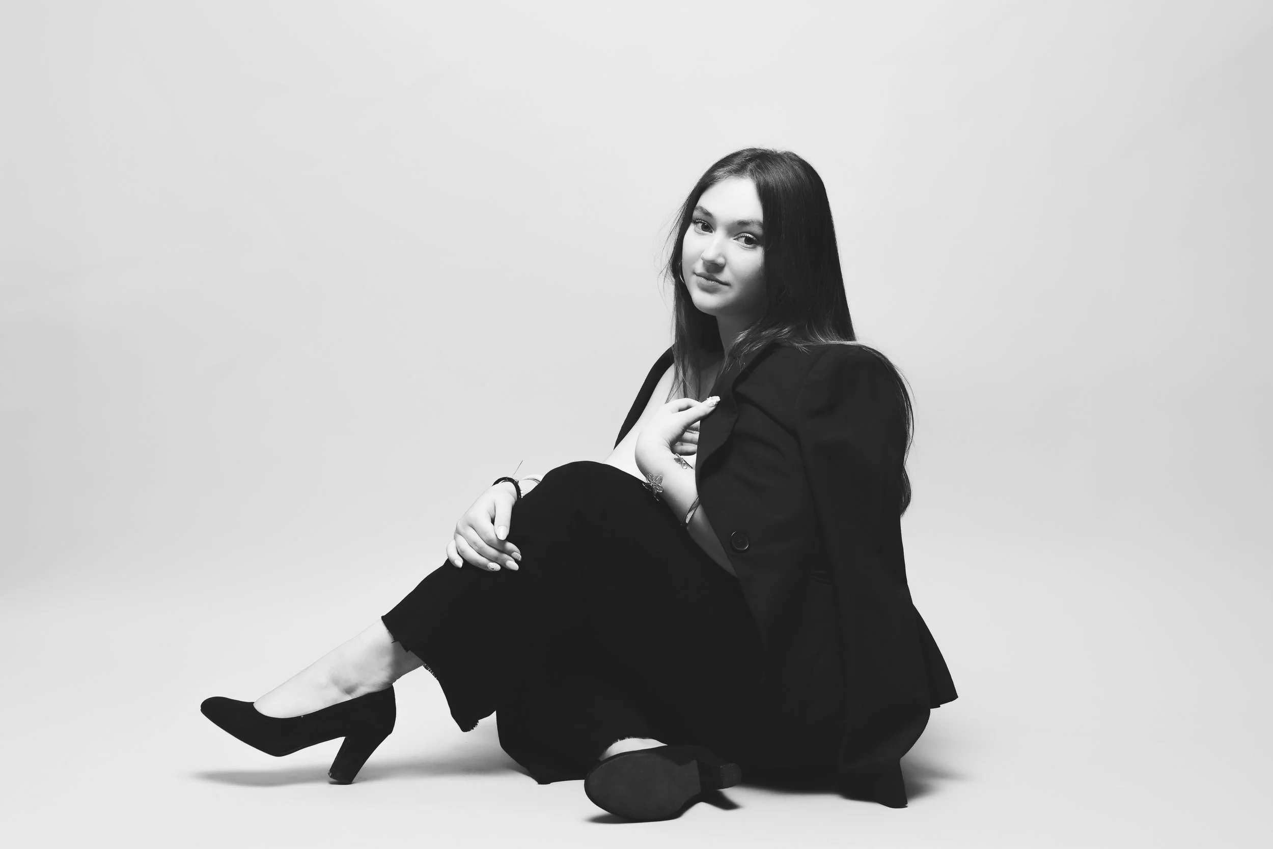 Une jeune femme aux cheveux longs, vêtue d'un costume noir, assise sur un siège, regardant vers l'appareil photo, dans un environnement minimaliste monochrome.