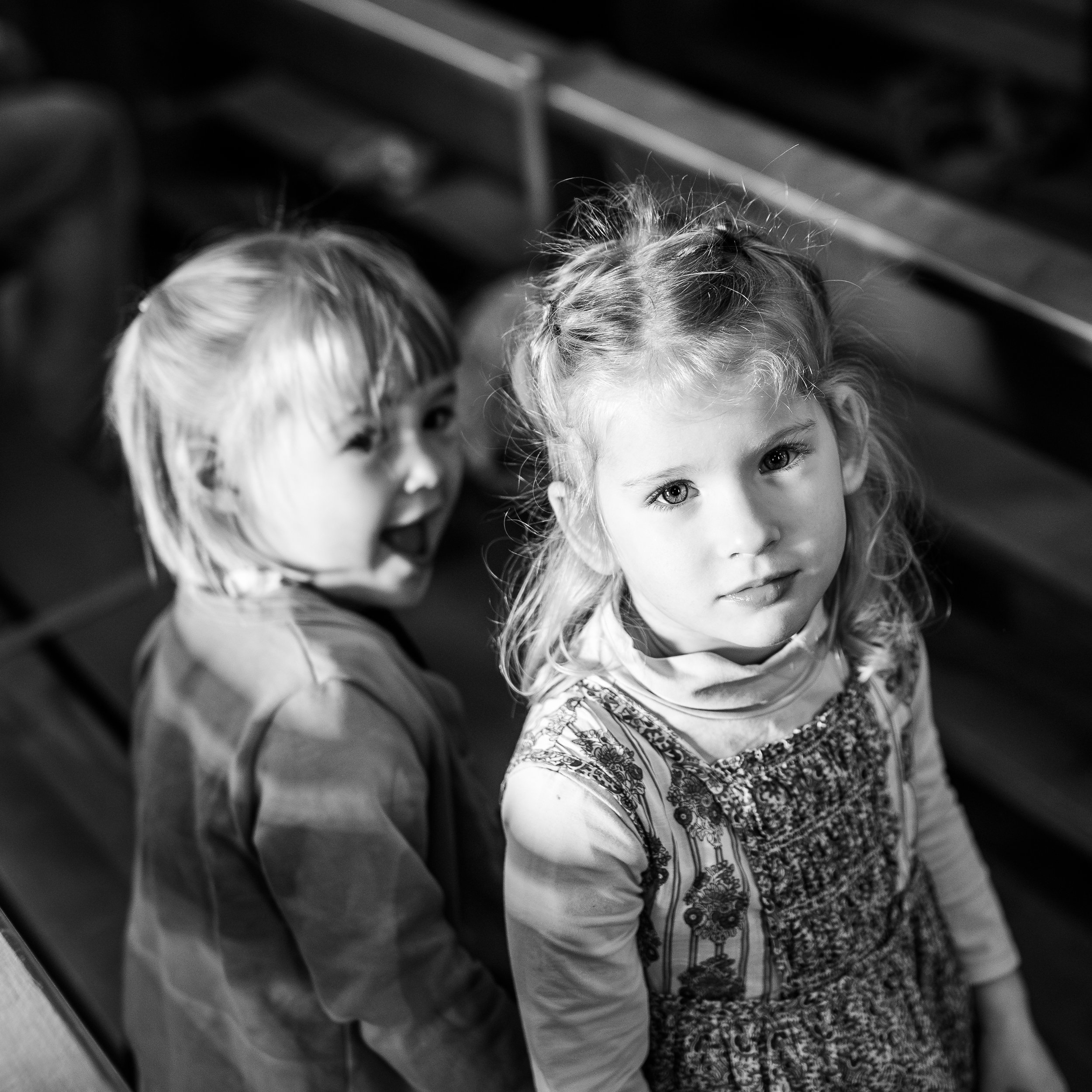 Deux jeunes filles avec des cheveux clairs, l'une a des yeux marron et l'autre des yeux bleus, regardant vers l'objectif, debout près d'une surface en marbre avec un fond en bois. L'image est en noir et blanc.