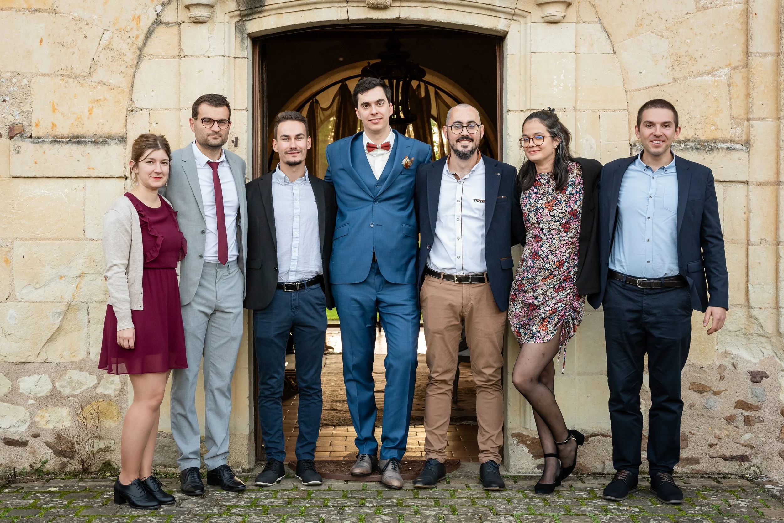 groupe de sept personnes habillées en vêtements formels, posant devant un bâtiment en pierre avec une arche en bois
