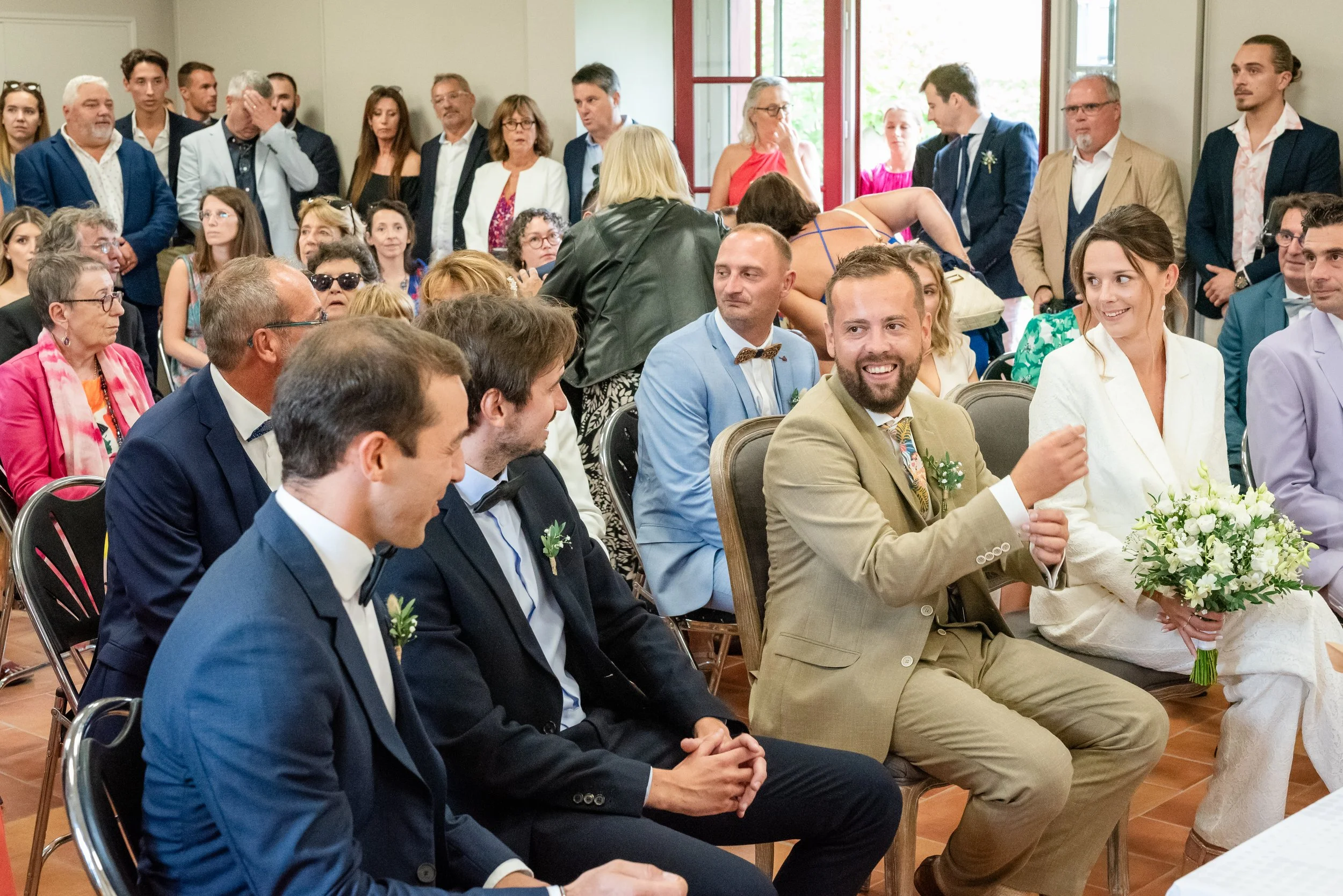 Un groupe de personnes lors d'une cérémonie ou d'une réception, beaucoup d'hommes en costume, une femme tenant un bouquet de fleurs blanches, dans une salle lumineuse avec fenêtres.