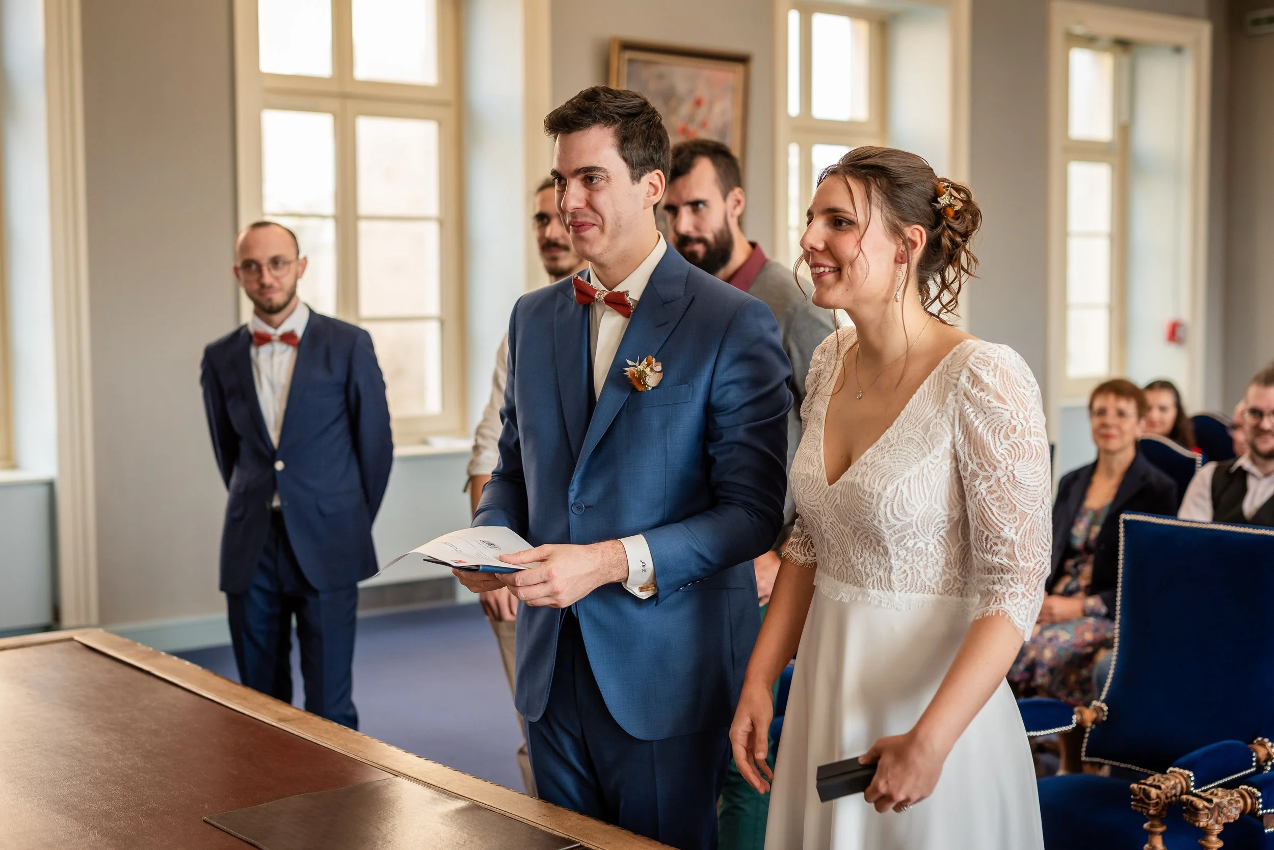 Un couple assiste à une cérémonie de mariage en intérieur, entouré de plusieurs témoins et invités, avec beaucoup de fenêtres lumineuses en arrière-plan.