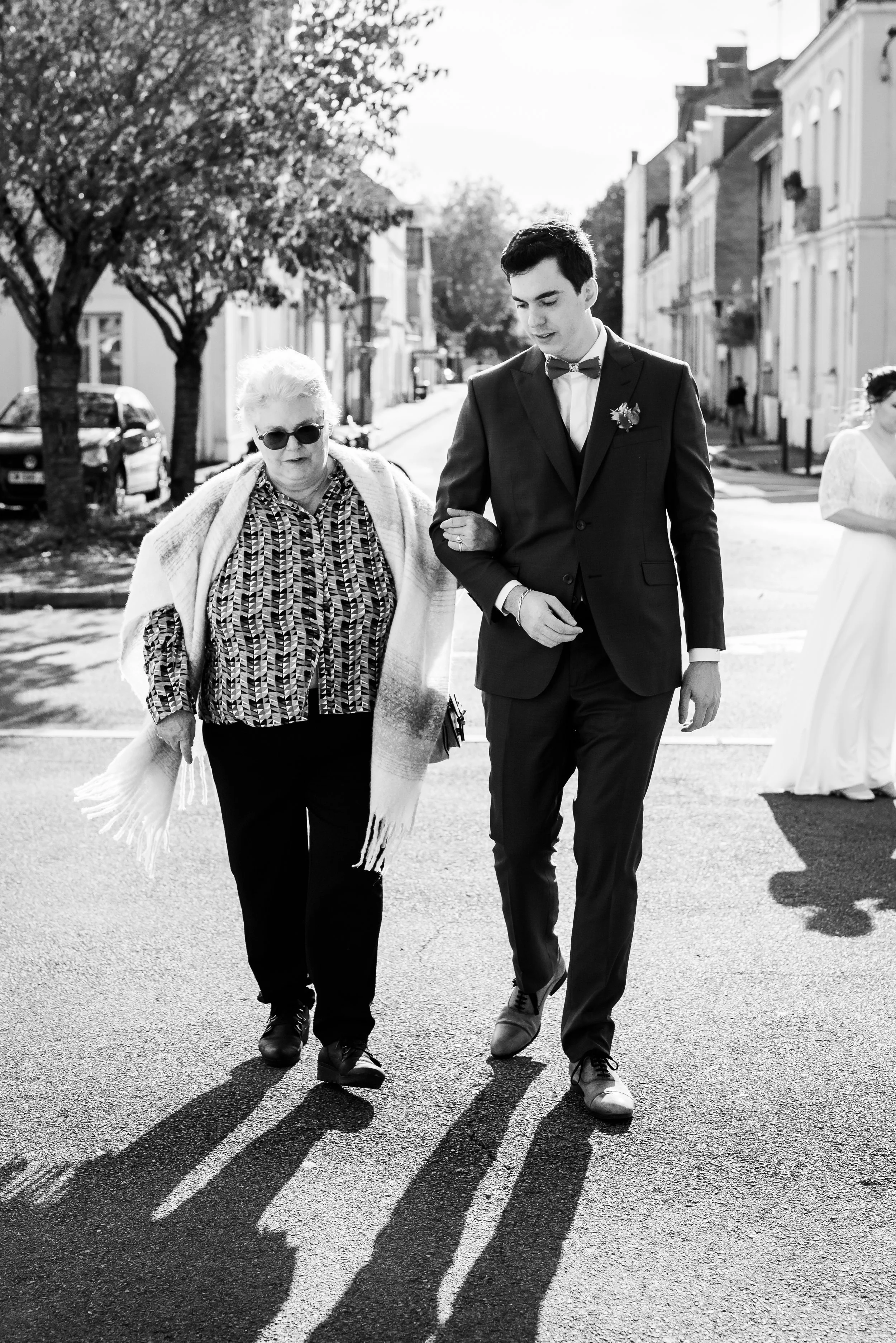 Deux personnes marchent côte à côte sur une rue, une femme âgée portant des lunettes et un manteau, et un jeune homme en costume avec une chemise et une cravate, plusieurs silhouettes de personnes en arrière-plan, arbres et maisons dans le quartier.