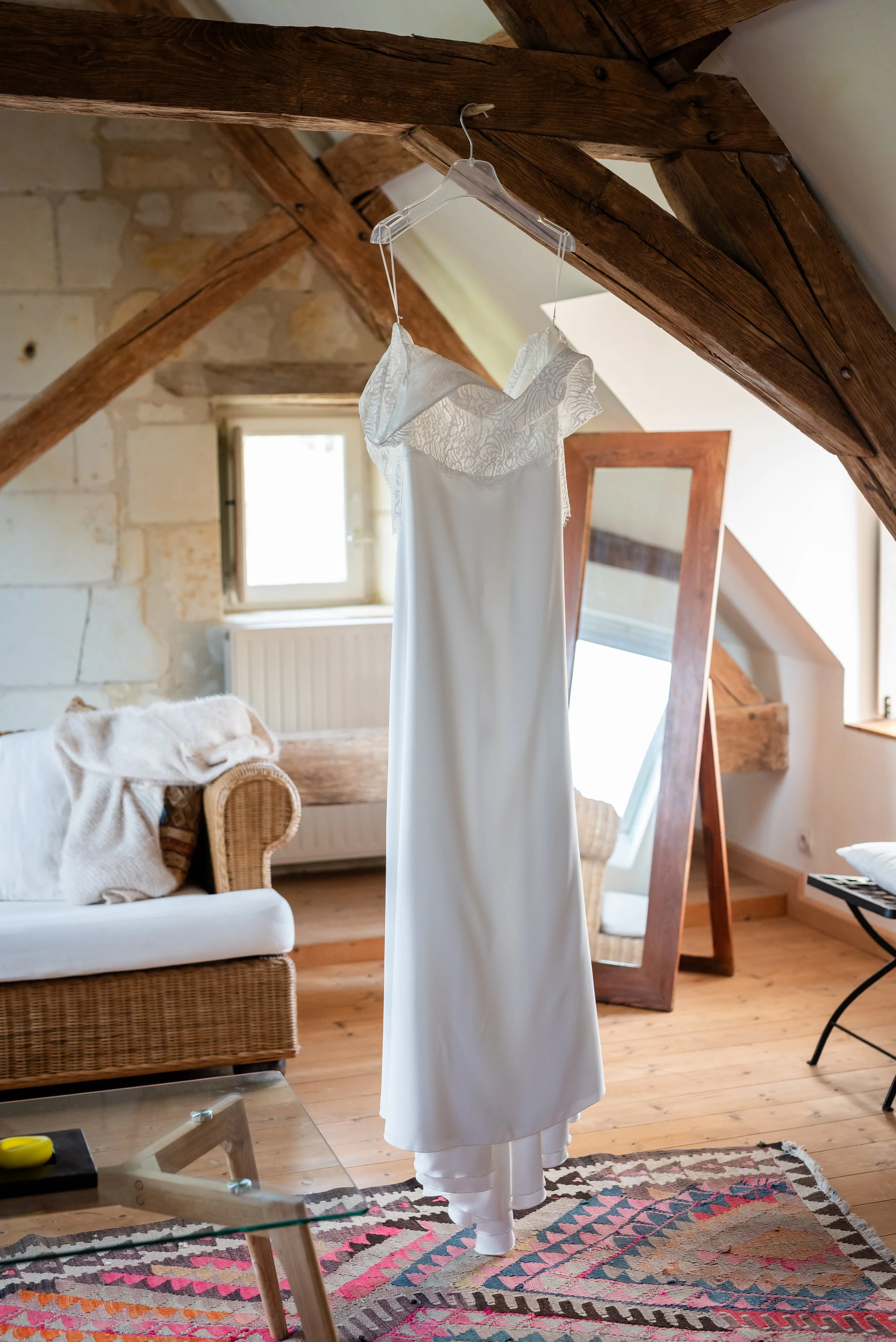 Une robe blanche suspendue à un cintre en plastique, accrochée à une poutre en bois dans une pièce avec un plafond en pente et un mur en pierre. La pièce possède une fenêtre et un miroir rectangulaire en bois. Il y a un canapé en rotin avec des couss