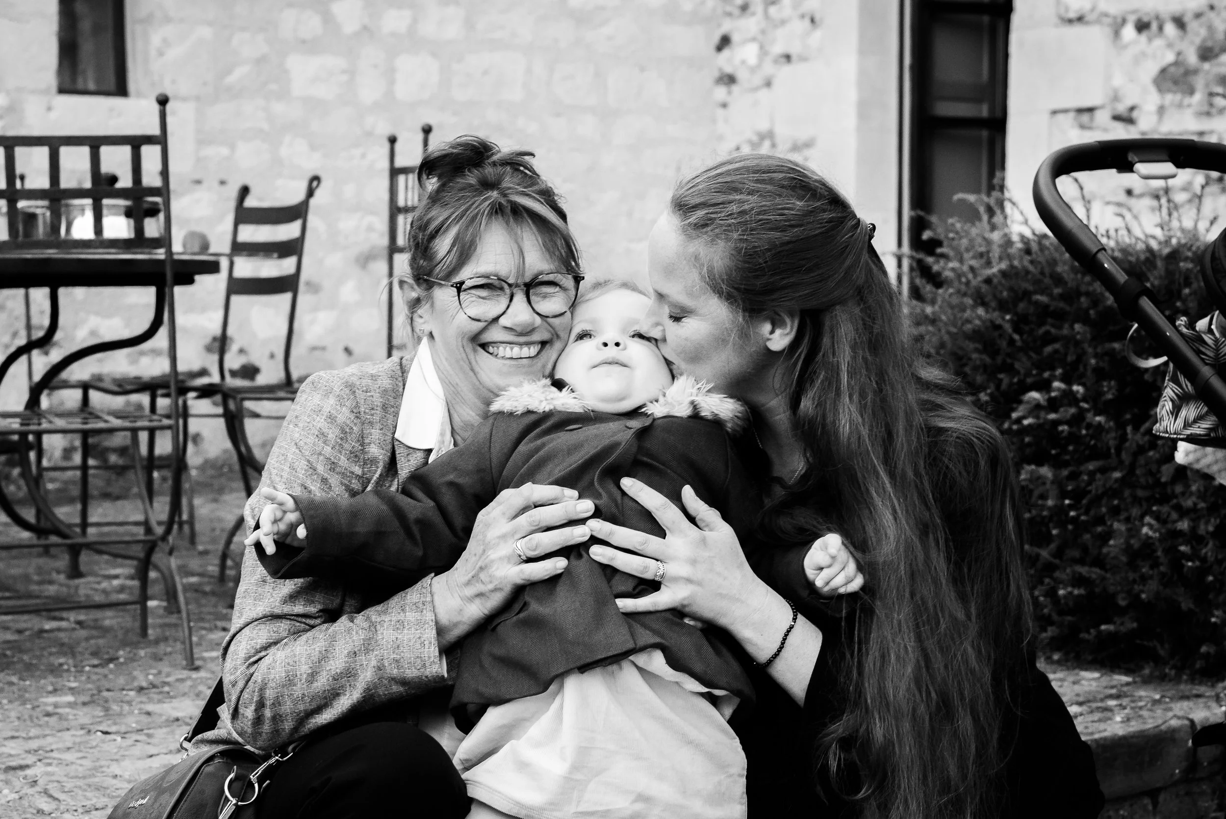Deux femmes et un bébé en plein câlin dans un espace extérieur avec des chaises en métal et un mur en pierre en arrière-plan.