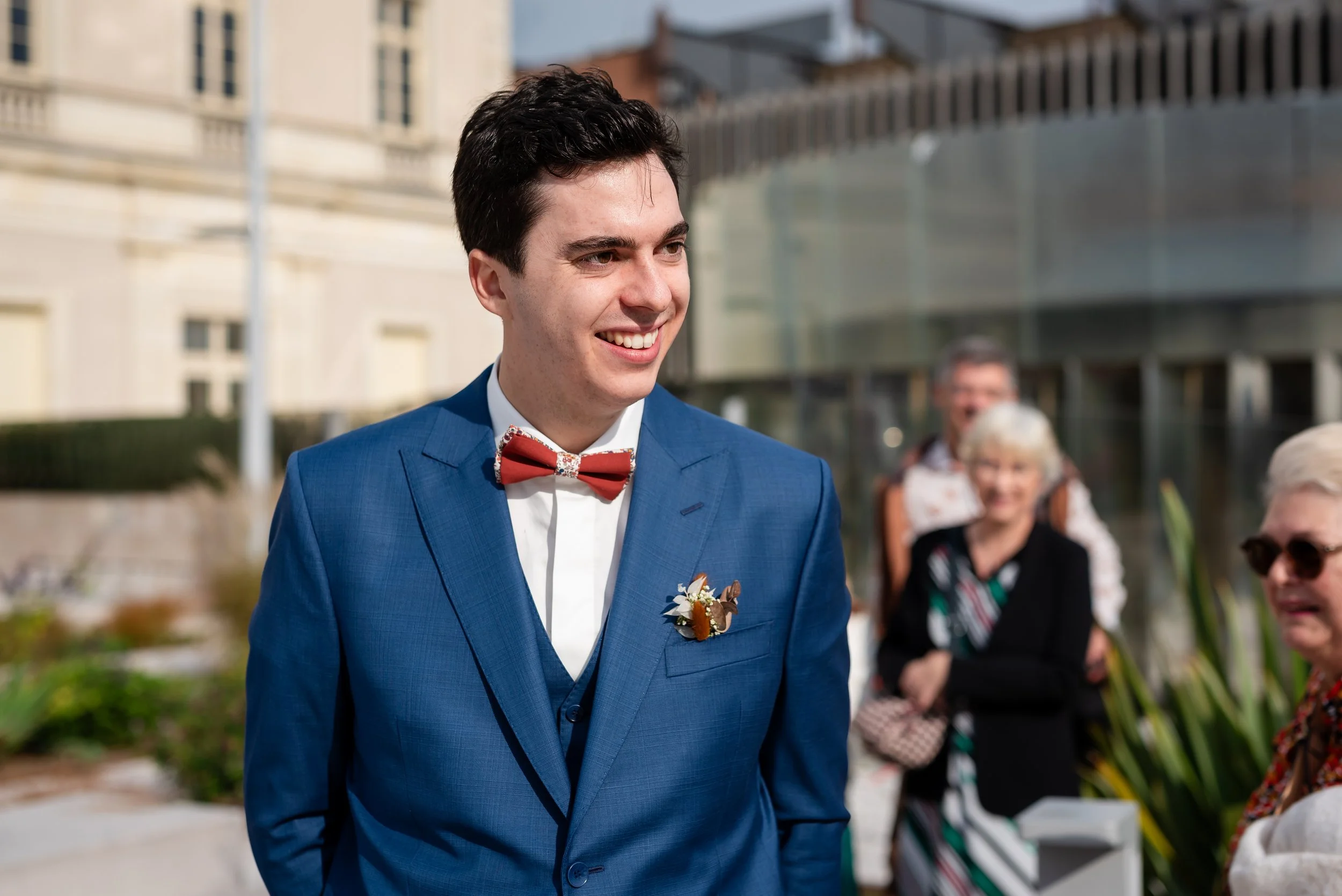 Un homme souriant porte un costume bleu avec un nœud papillon rouge lors d'une célébration extérieure, entouré de personnes âgées.