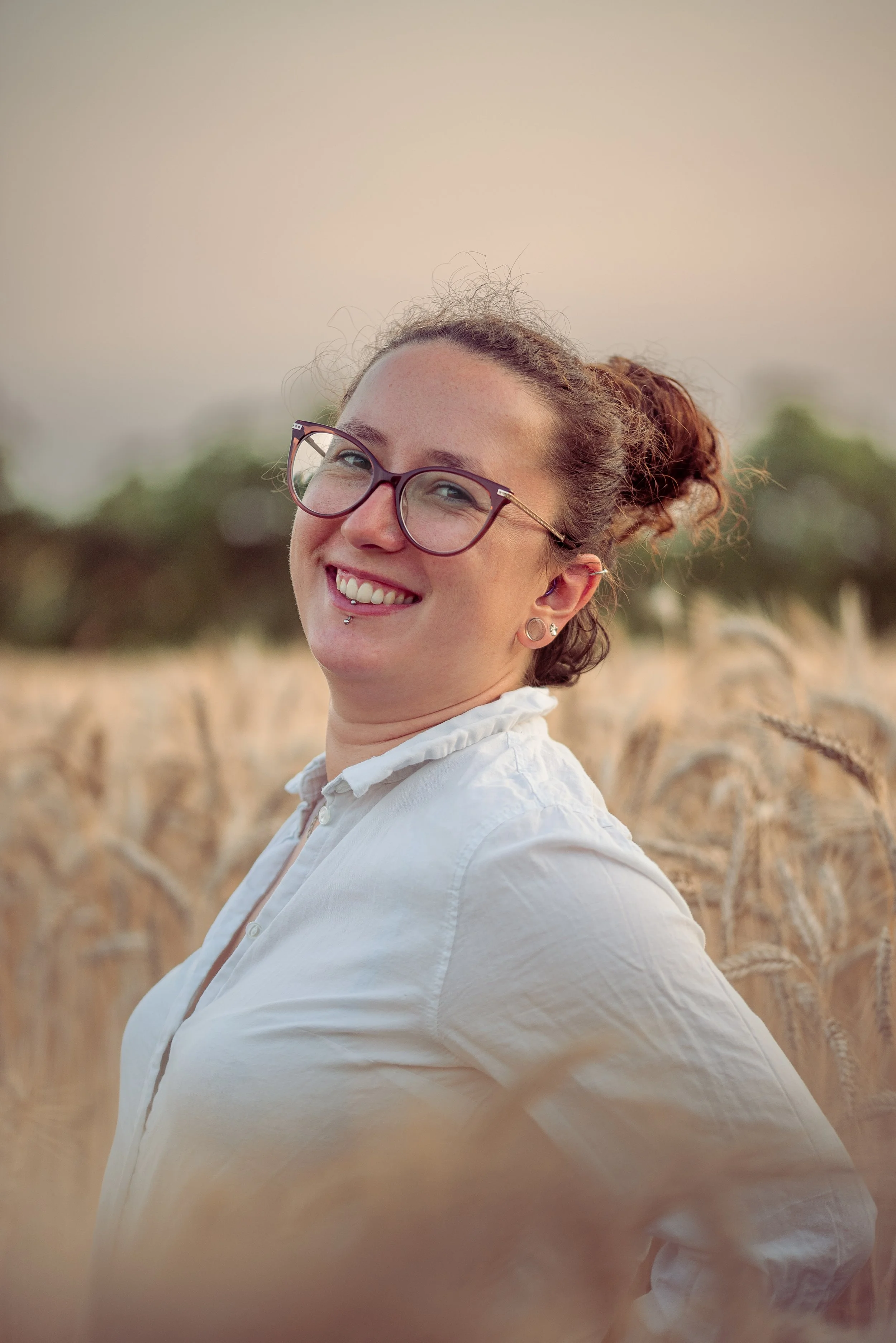 Jeune femme souriante avec lunettes, piercing au labret, dans un champ de blé, portant une chemise blanche.