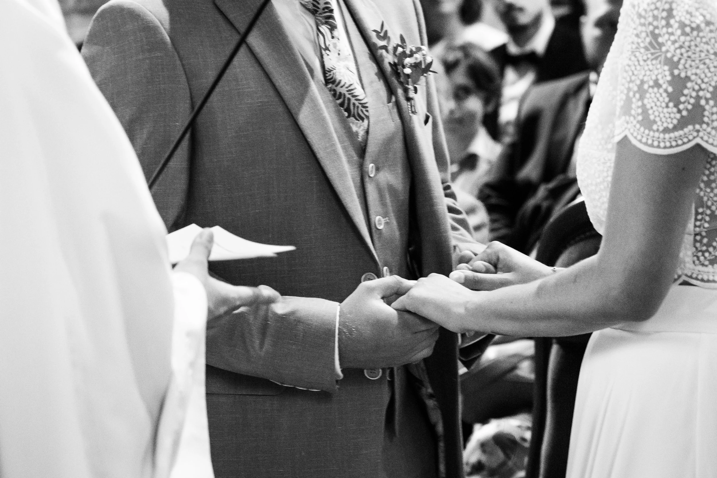 Une scène en noir et blanc d'une cérémonie, probablement un mariage, où deux personnes échangent un anneau, entourées d'autres personnes. La personne au centre porte un costume avec une boutonnière.