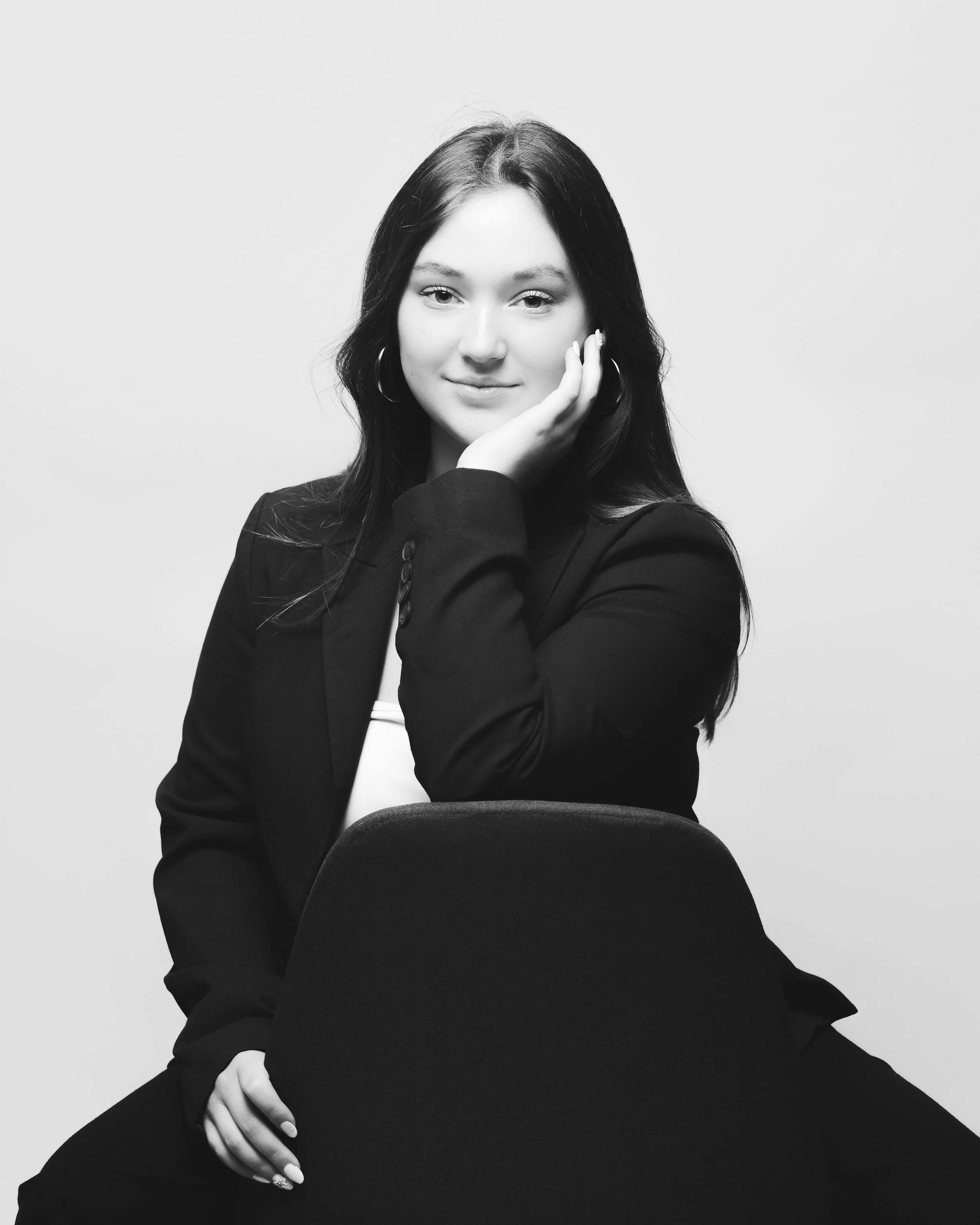 Portrait en noir et blanc d'une femme souriante avec les cheveux longs et foncés, portant un blazer noir et un top blanc, posant avec la main sur la joue.