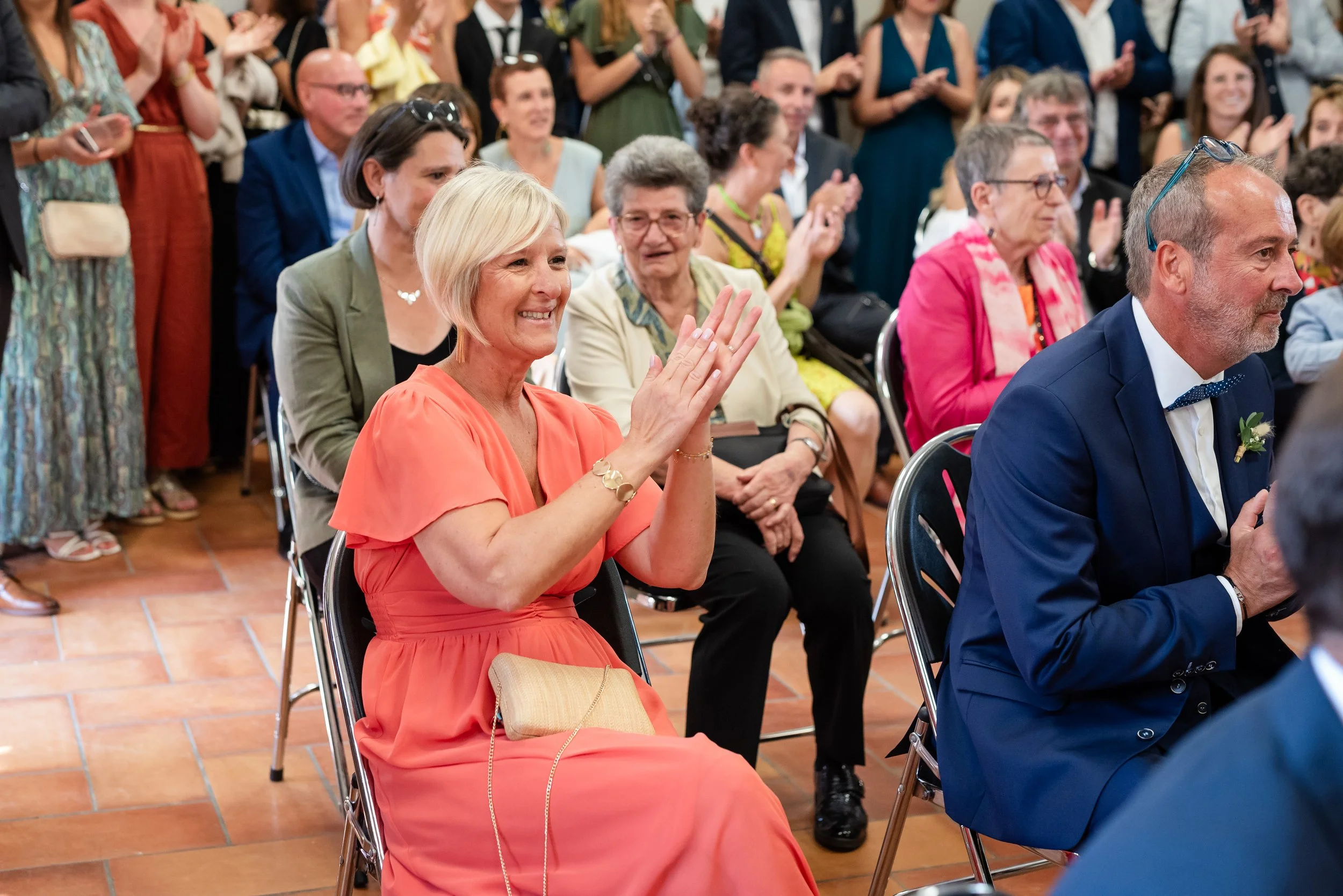 Un groupe de personnes assises en train d'assister à un événement, applaudissant et souriant, avec une femme en premier plan portant une robe habillée en orange, qui sourit et applaudit.