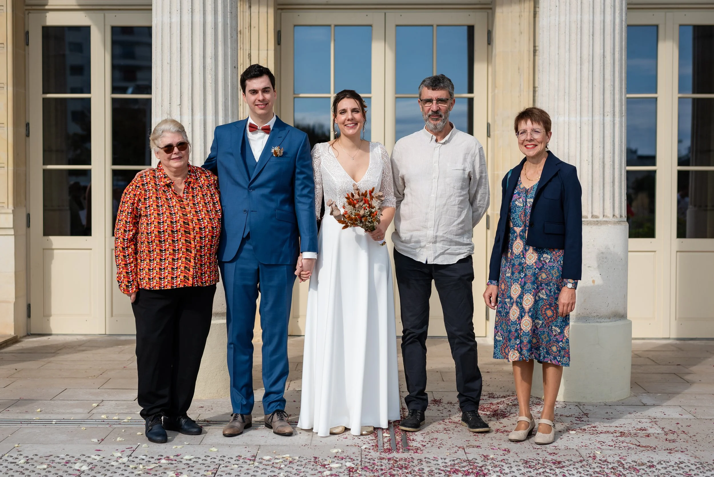 Cinq personnes posent devant un bâtiment lors d'un mariage. La mariée tient un bouquet de fleurs. La scène semble festive avec des pétales de fleurs sur le sol.