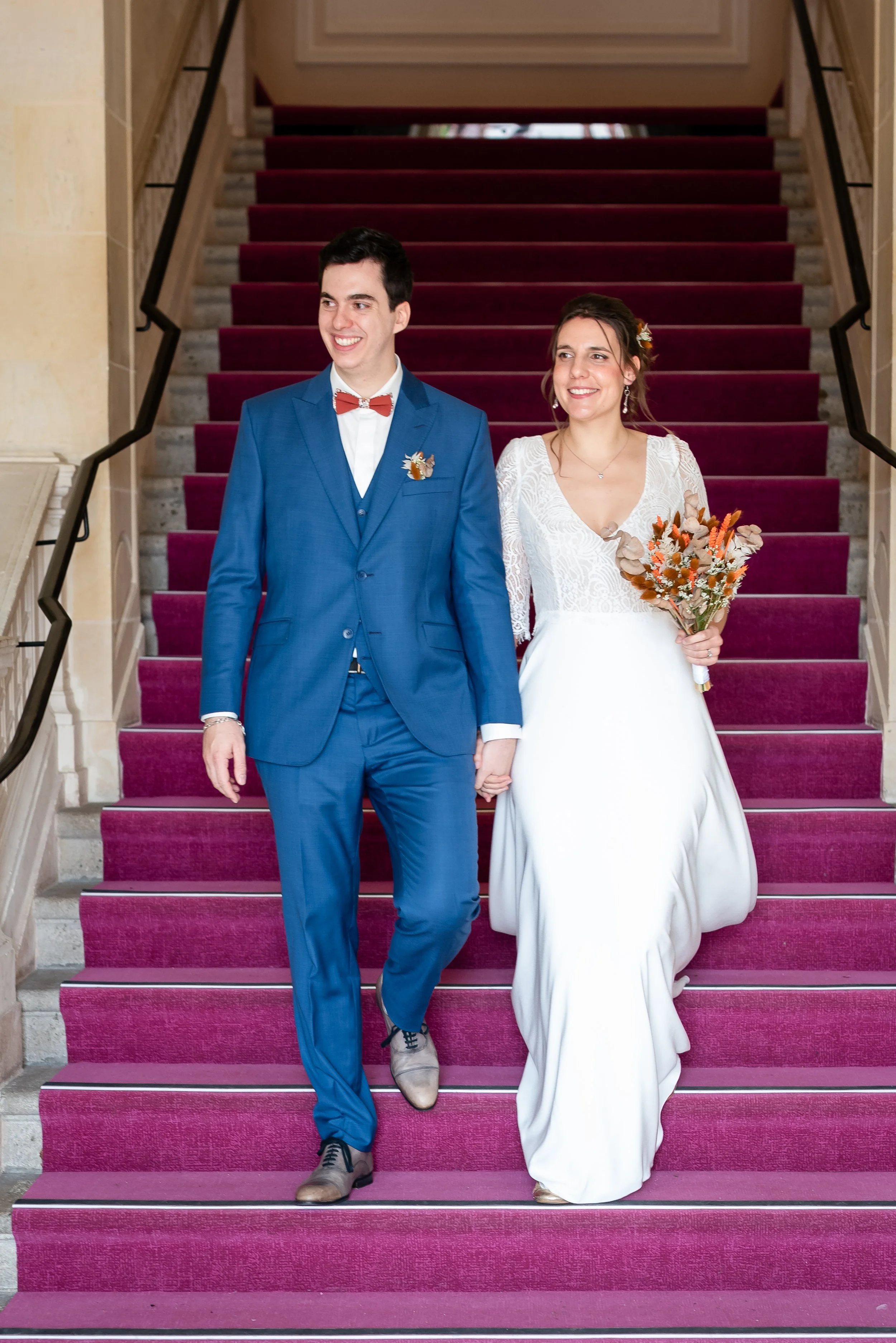 Un couple en mariage descend un escalier avec tapis rouge, la femme porte une robe blanche et tient un bouquet, l'homme porte un costume bleu avec nœud papillon rouge.