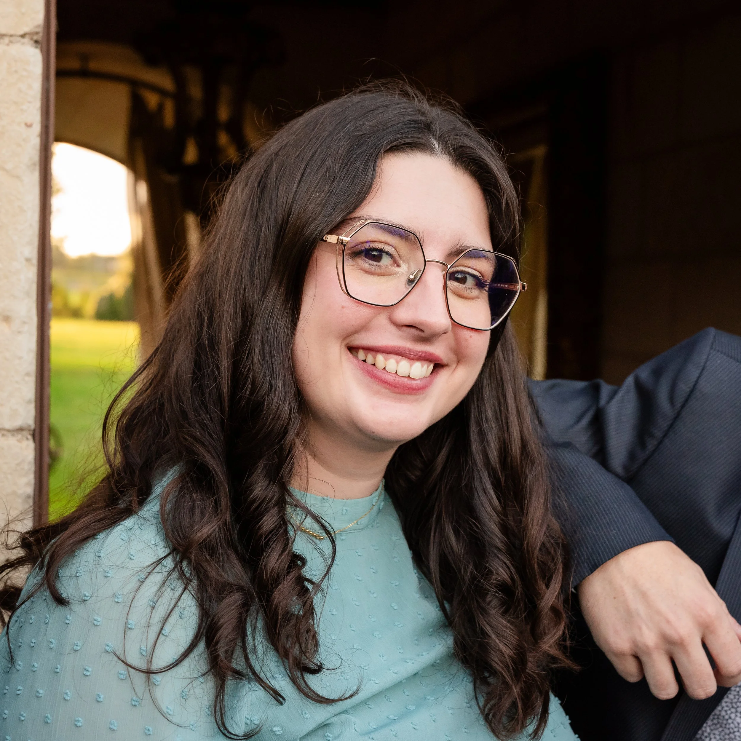 Jeune femme souriante avec des lunettes, cheveux longs et brun foncé, portant une chemise bleue avec des motifs en relief, prise en photo lors d'une occasion sociale extérieure.