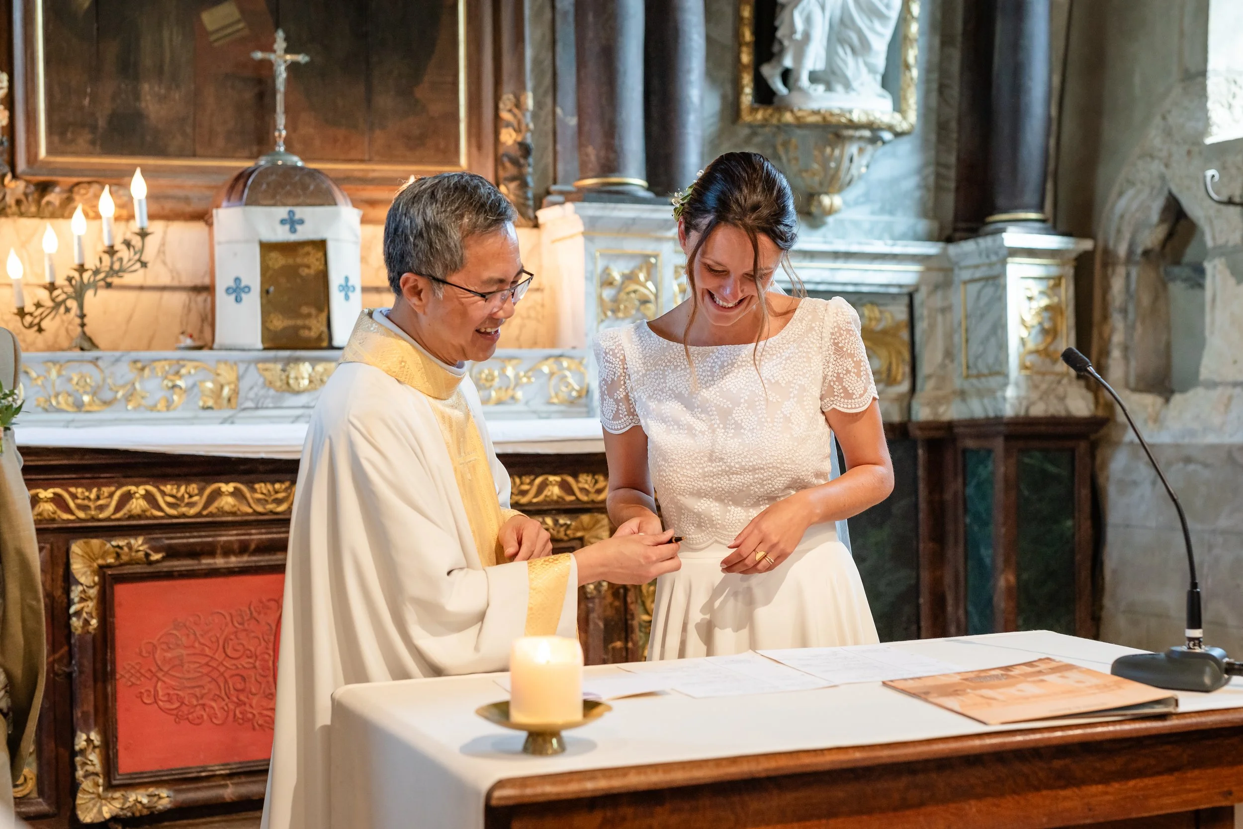 Une cérémonie religieuse, probablement un mariage, dans une église ancienne. Un prêtre en robe blanche et or, un homme et une femme souriant, se tenant par la main lors de l'échange des vœux ou d'une bague, devant un autel orné de bougies et d'une cr