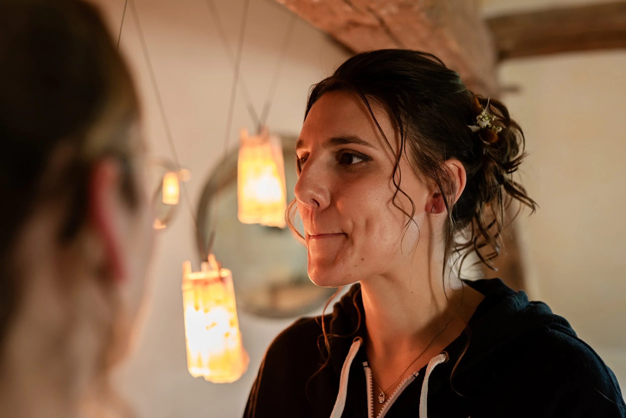 Une jeune femme se regarde dans un miroir, l'air concentrée, dans un environnement chaleureux avec des lampes suspendues en arrière-plan.