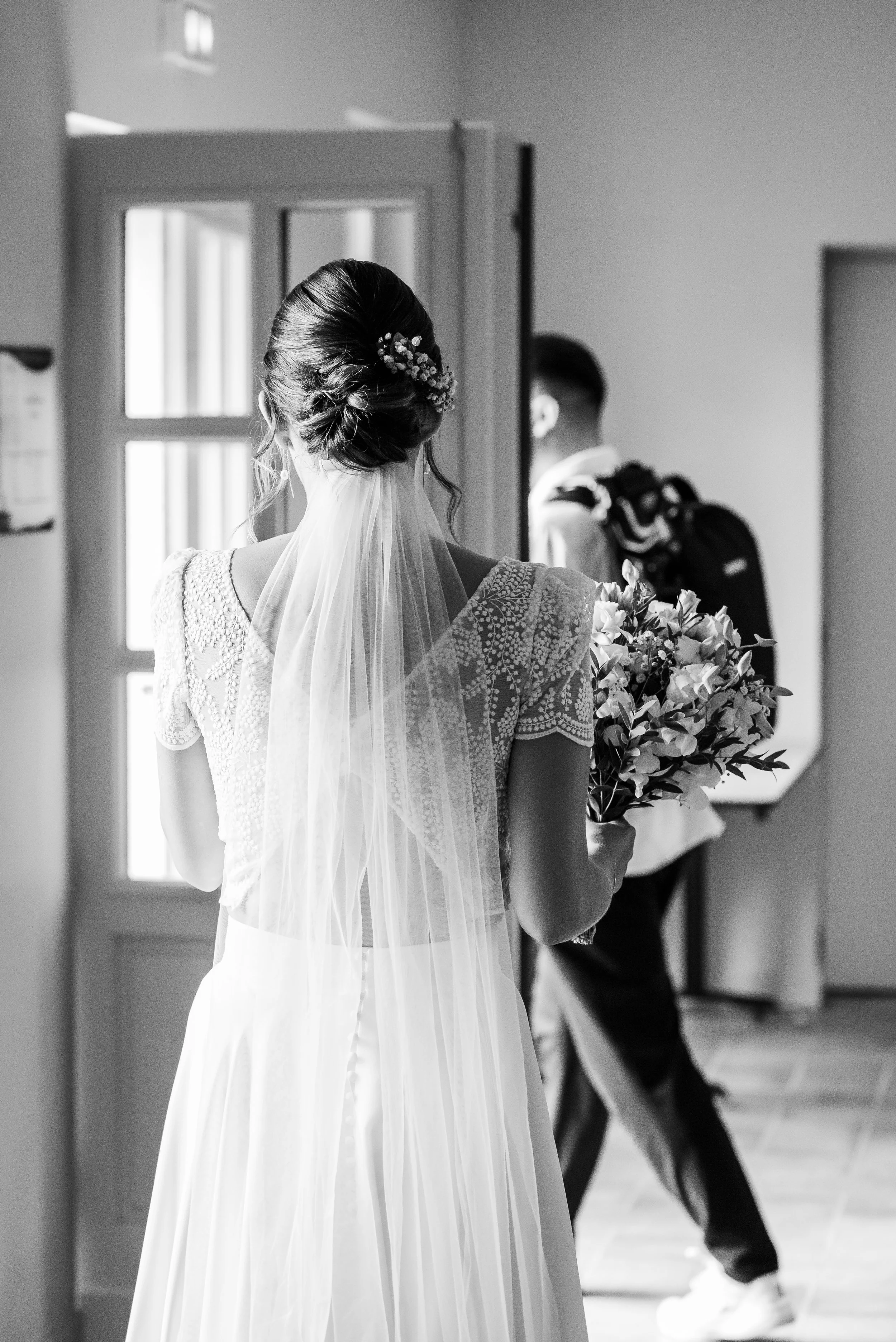 Une photo en noir et blanc d'une femme vêtue d'une robe de mariée avec un voile, vue de dos, tenant un bouquet de fleurs, dans un intérieur. En arrière-plan, un homme en tenue décontractée porte un sac à dos.
