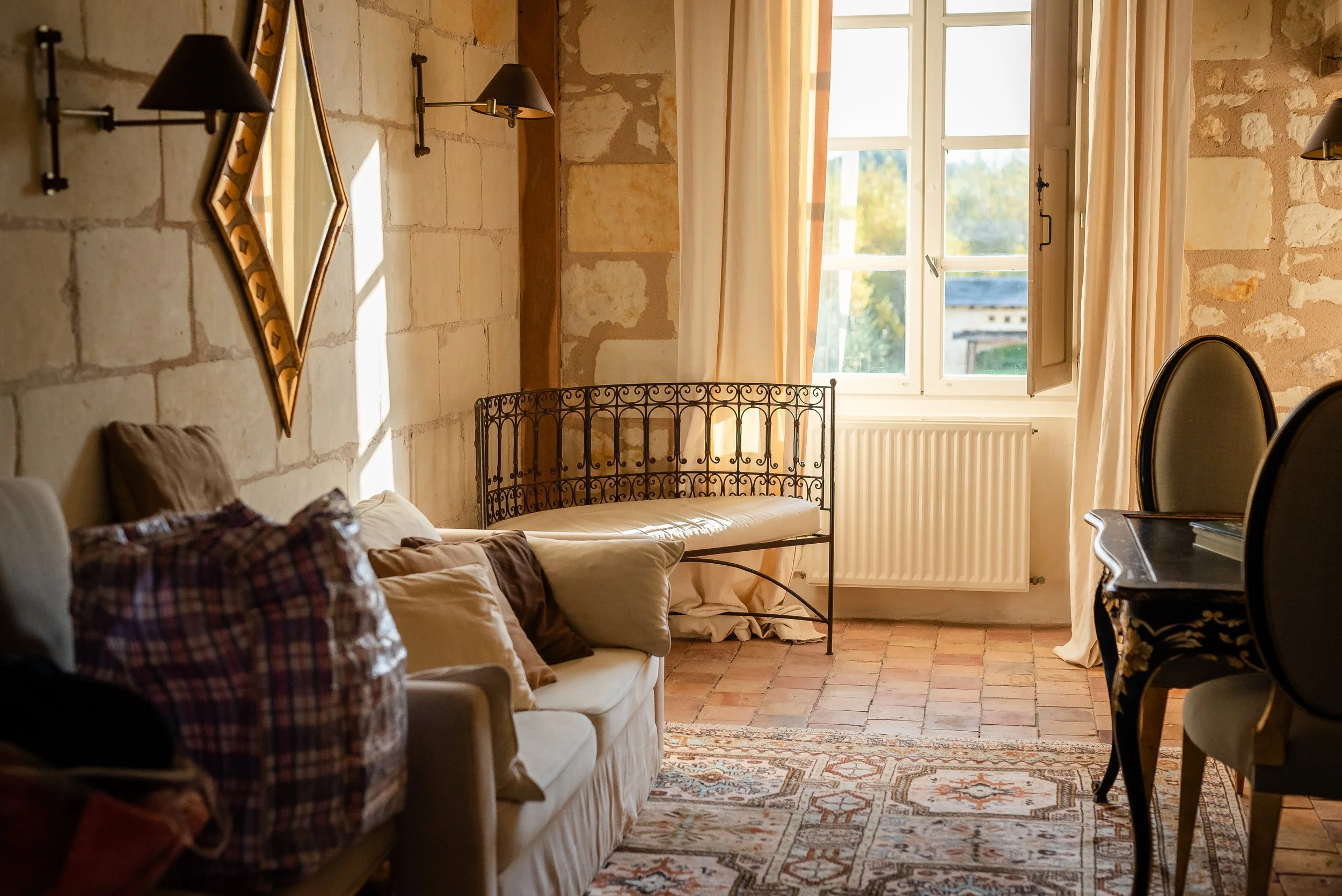 Salon avec canapé, coussins, fauteuils, grande fenêtre avec rideaux, décor en pierre, tapis oriental, mur avec miroir et lampes