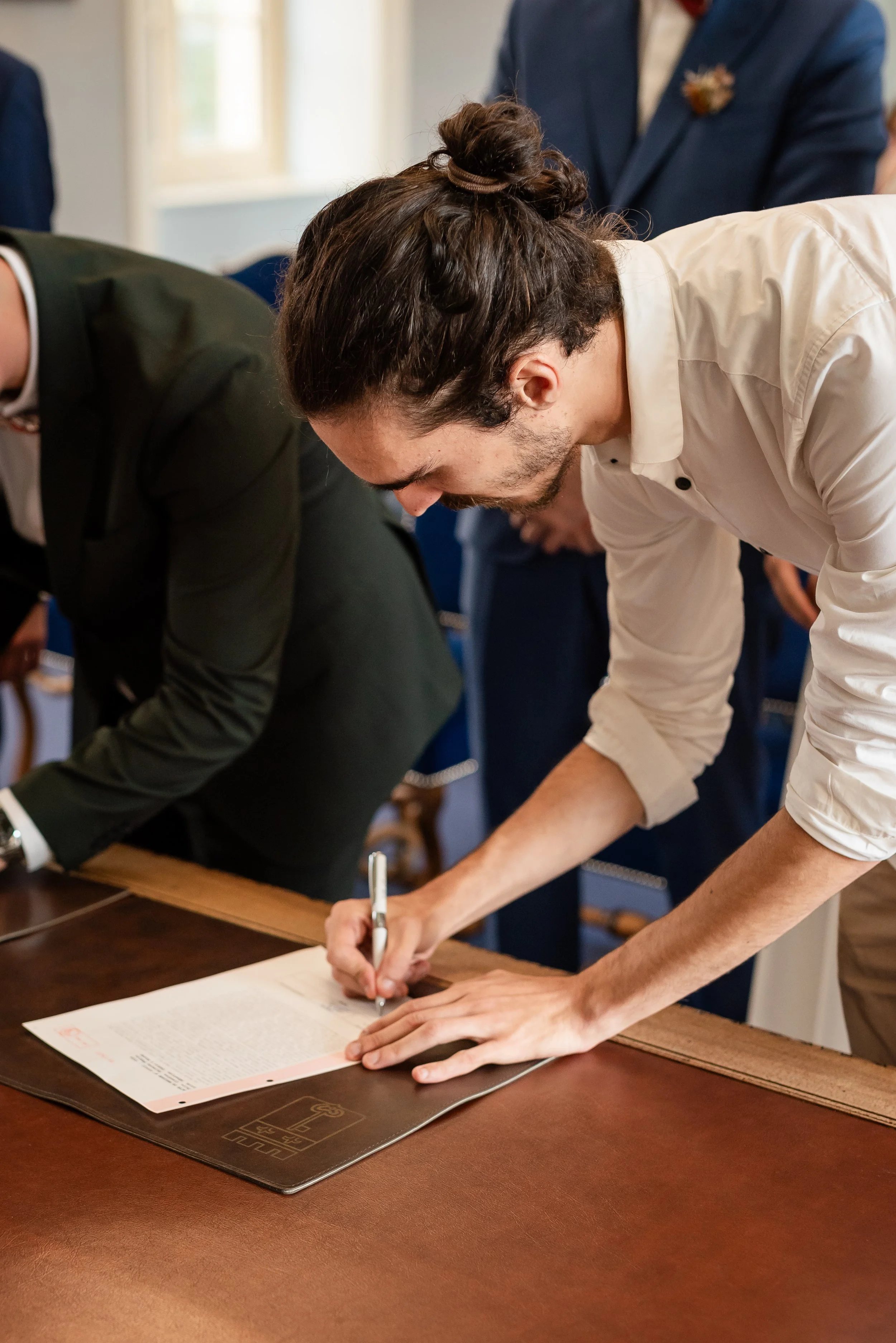 Deux hommes signent un document sur une table en bois dans une salle éclairée par la lumière naturelle.