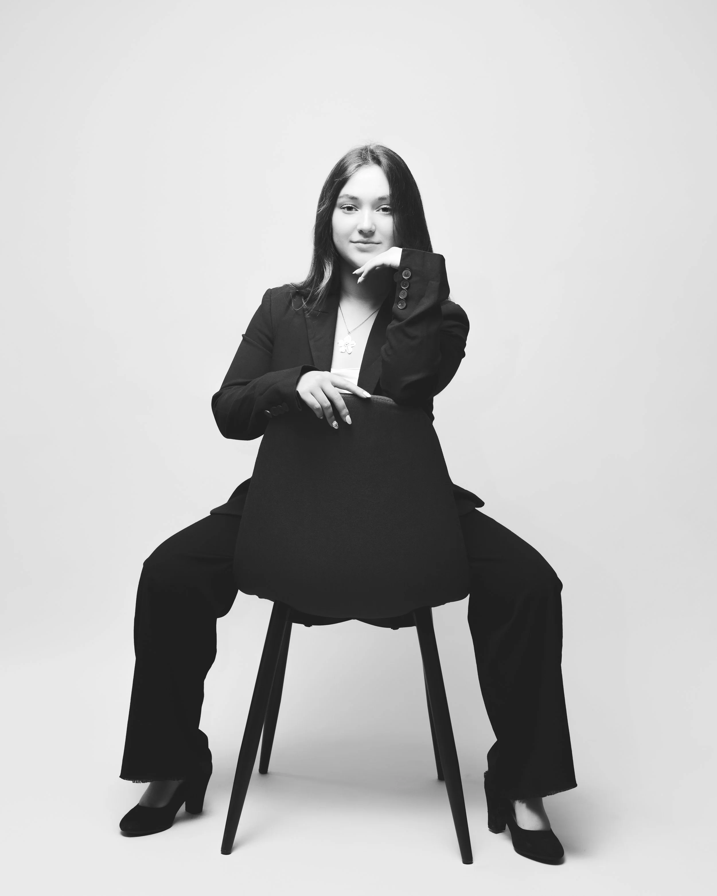Une femme en noir et blanc, habillée d'une veste noire, posant avec confiance tout en étant assise sur une chaise, les jambes écartées, dans un studio avec un fond blanc ou gris clair.