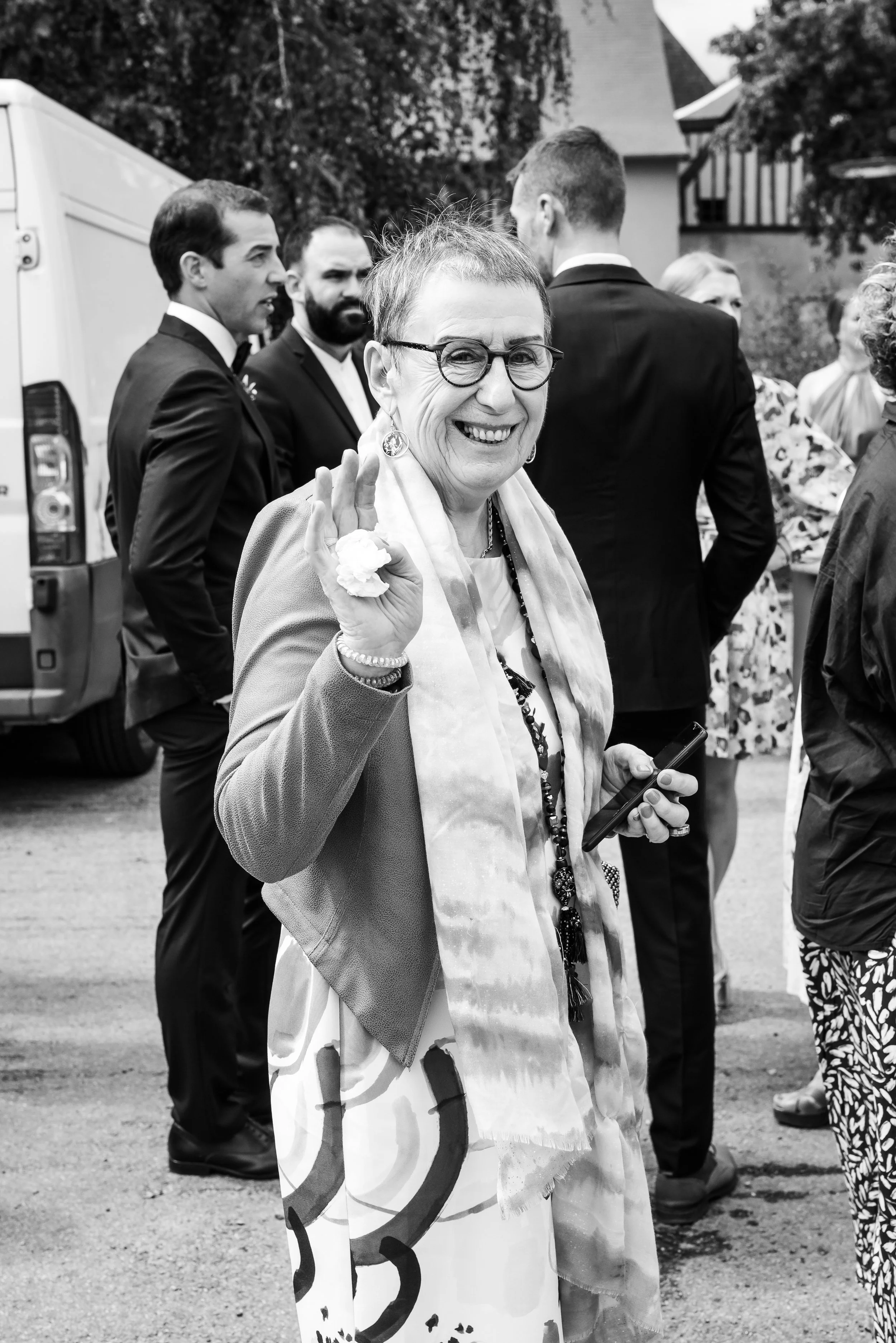 Une femme âgée souriante portant des lunettes et un sourire éclatant fait un signe de la main lors d'une réunion en plein air, entourée de plusieurs personnes en tenue formelle