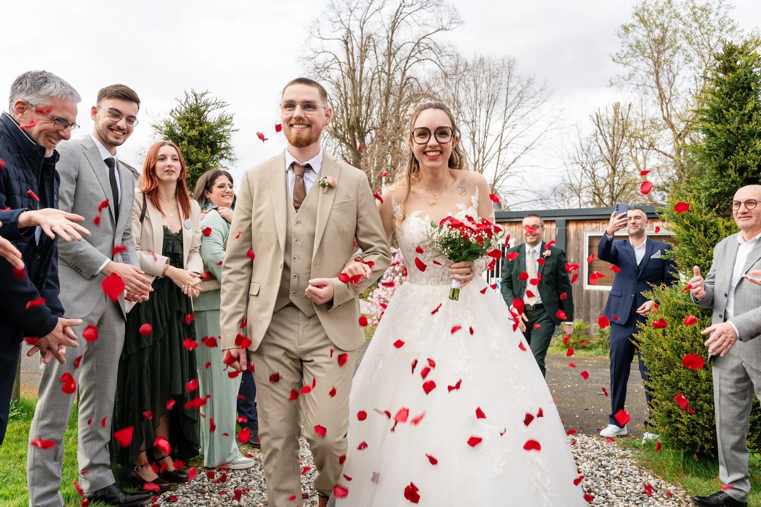 Un couple de mariés marchant sous des pétales rouges, entouré d'amis et de famille lors d'une cérémonie en plein air.