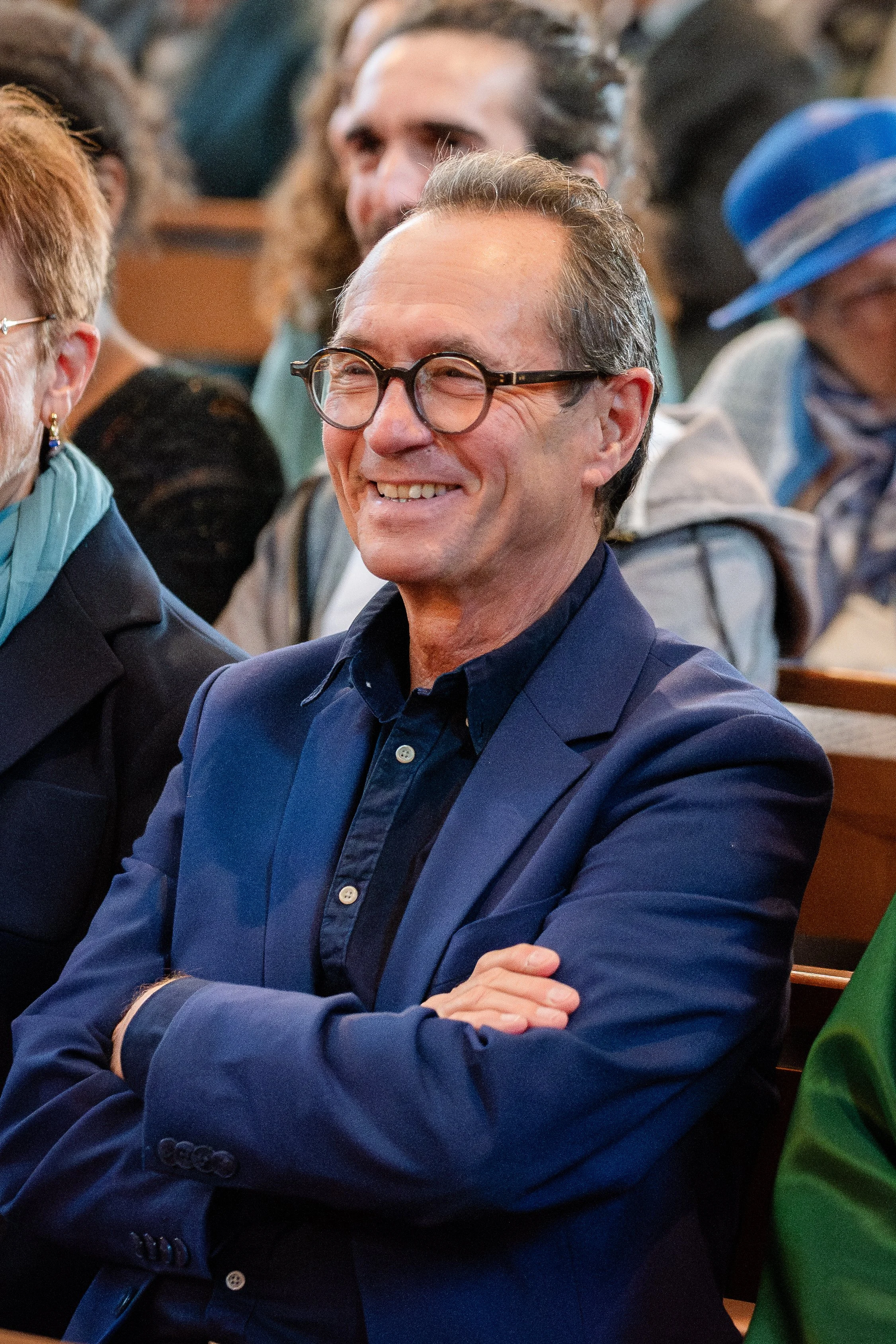 Un homme souriant portant des lunettes, habillé en chemise bleue, assis avec les bras croisés dans une salle remplie de personnes, probablement lors d'un événement ou d'une réunion.