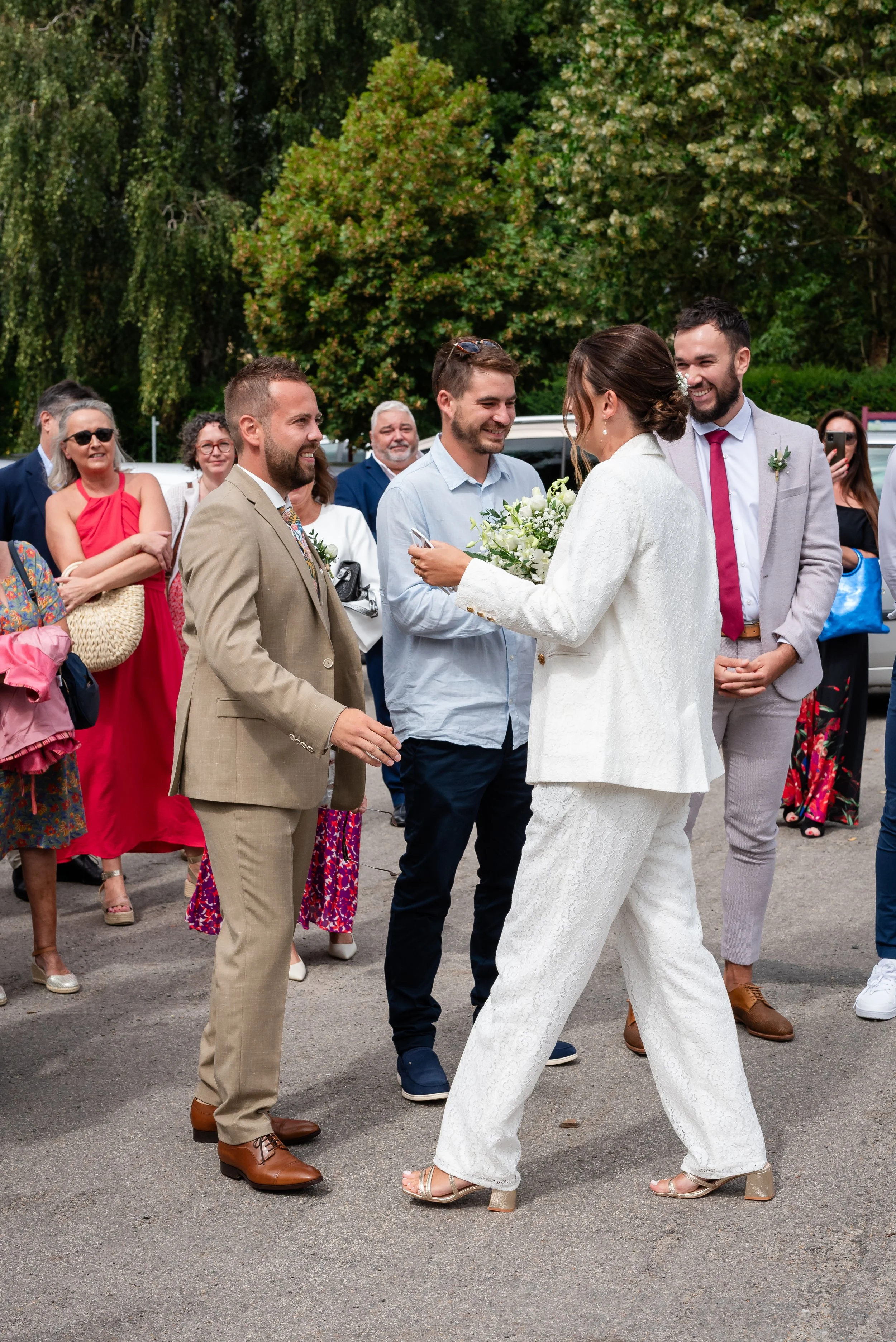 Un groupe de personnes au mariage, la mariée en robe blanche et les invités souriants, dans un espace extérieur avec des arbres en arrière-plan.