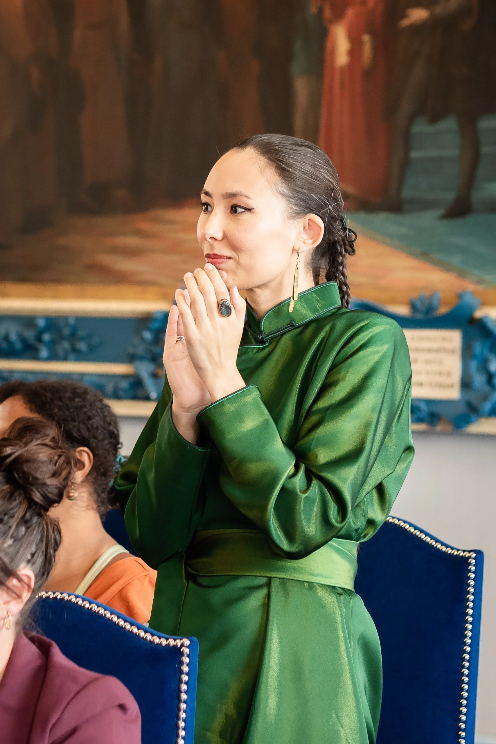 Une femme asiatique en robe verte, avec que ses mains jointes près du visage, semble prier ou méditer, dans un lieu décoré avec des œuvres d'art.