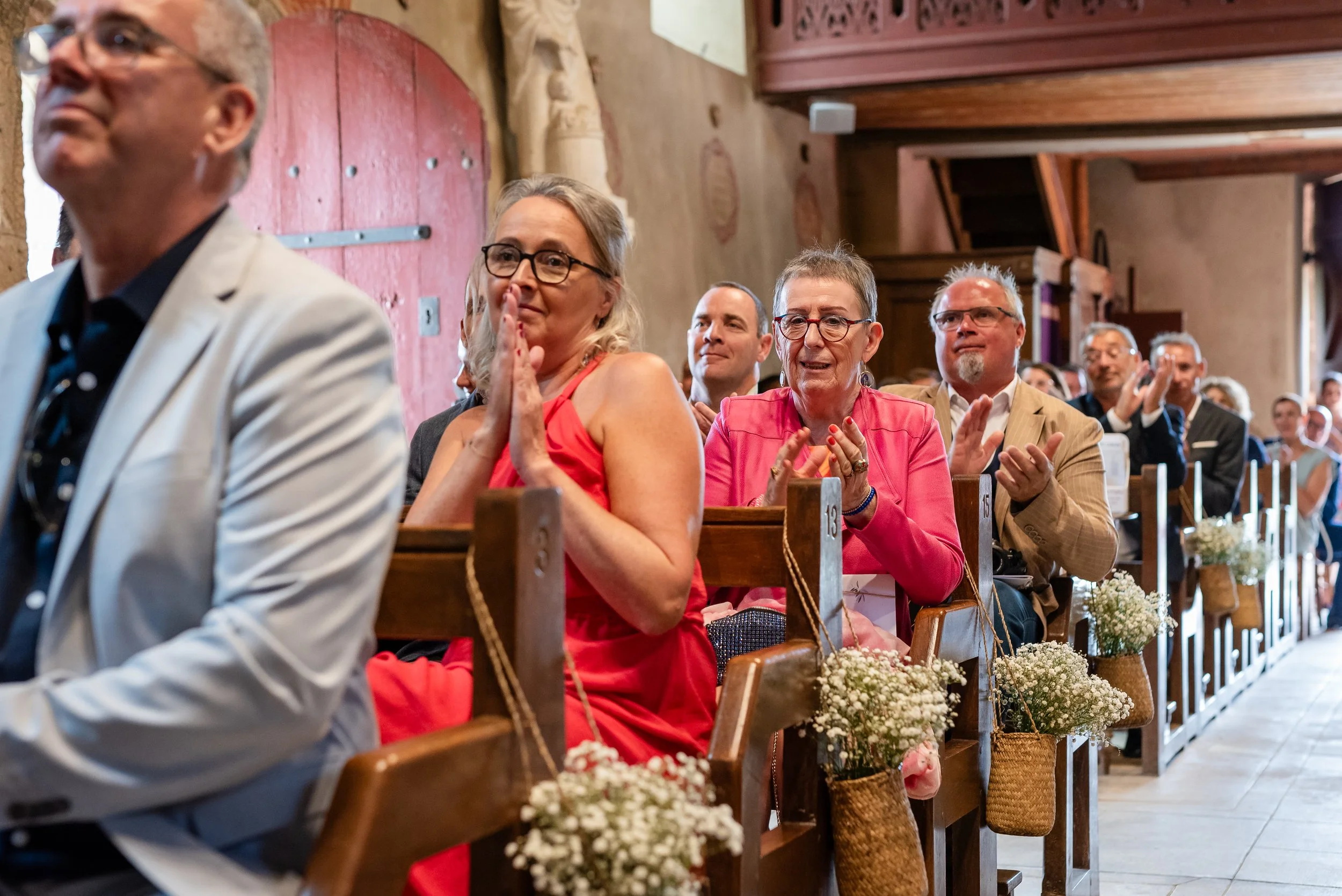 Groupe de personnes assises en rangée dans une salle décorée de fleurs, applaudissant lors d'une cérémonie ou d'un événement.