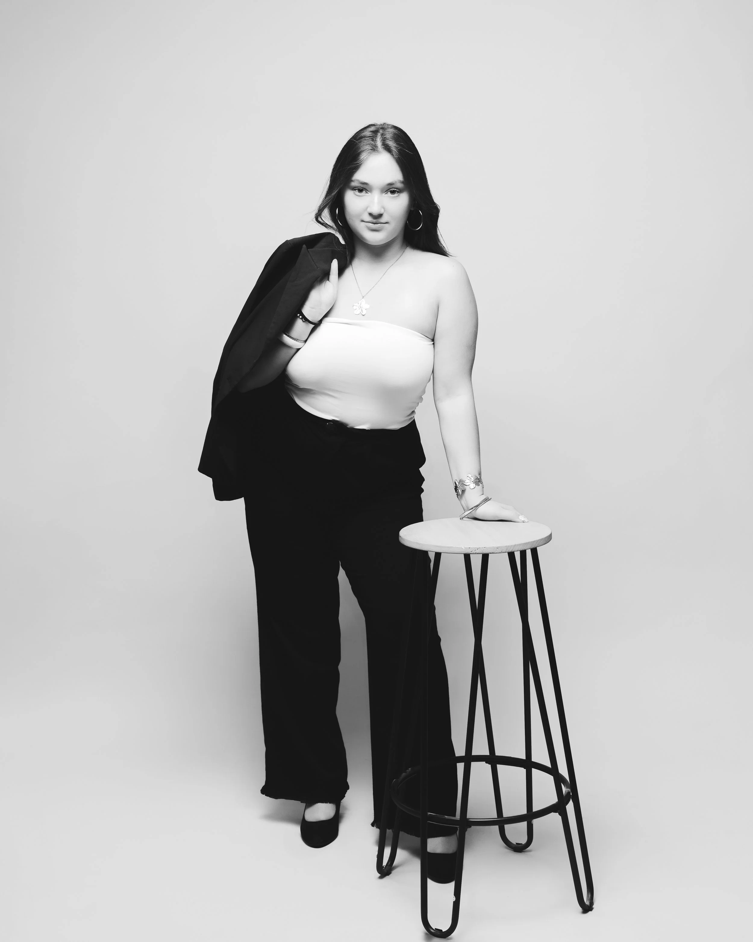 Une femme pose avec une veste sur son épaule, regardant vers l'objectif, en noir et blanc, devant un fond simple, avec un tabouret à côté d'elle.
