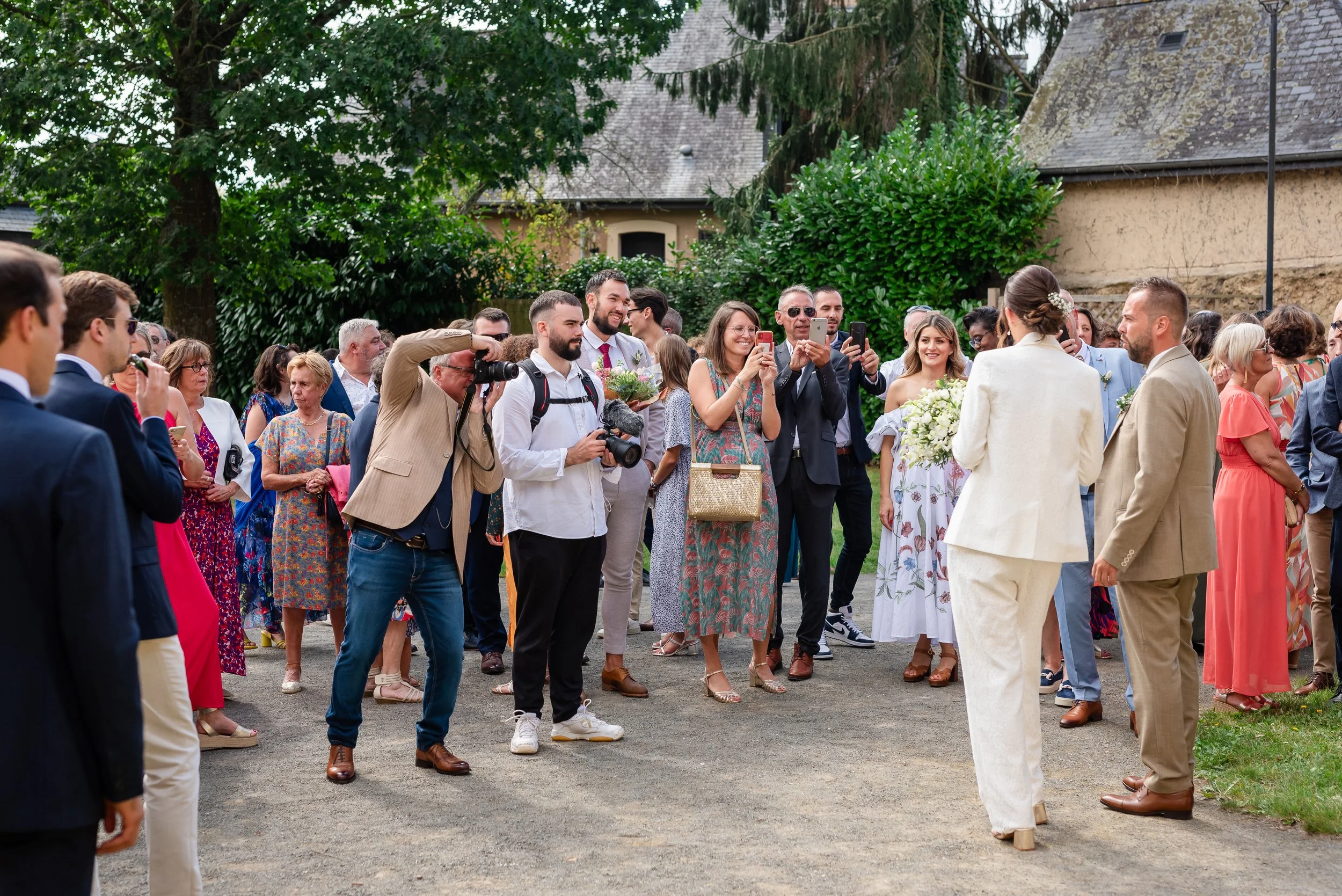 Groupe de personnes lors d'une cérémonie ou d'un mariage, la mariée en blanc et le marié en beige face à une assemblée, certains prennent des photos, dans un jardin avec des arbres et une vieille bâtisse en arrière-plan.