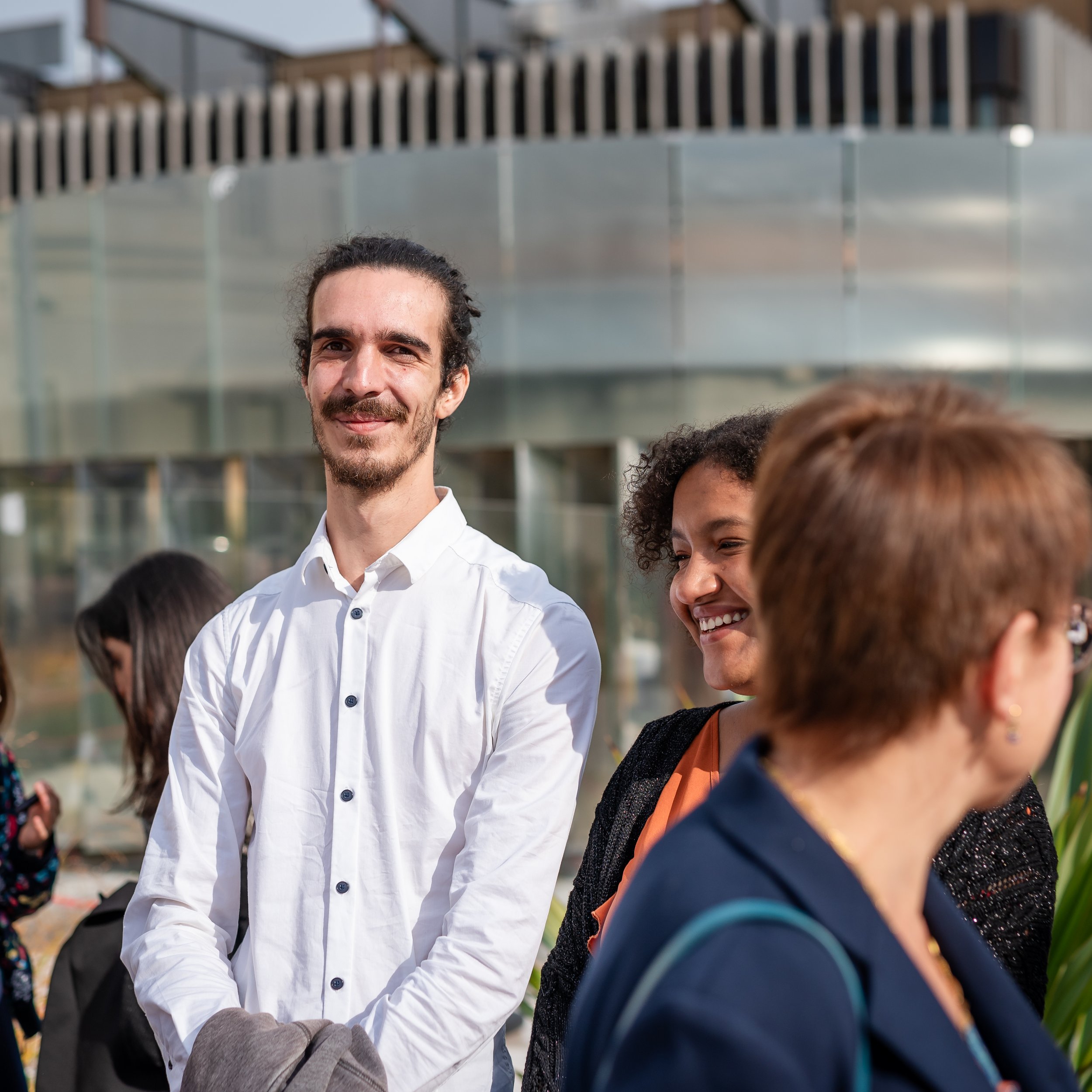 Groupe de personnes souriantes en extérieur, dont un homme avec une barbe et un sourire, portant une chemise blanche, entouré d'autres individus.