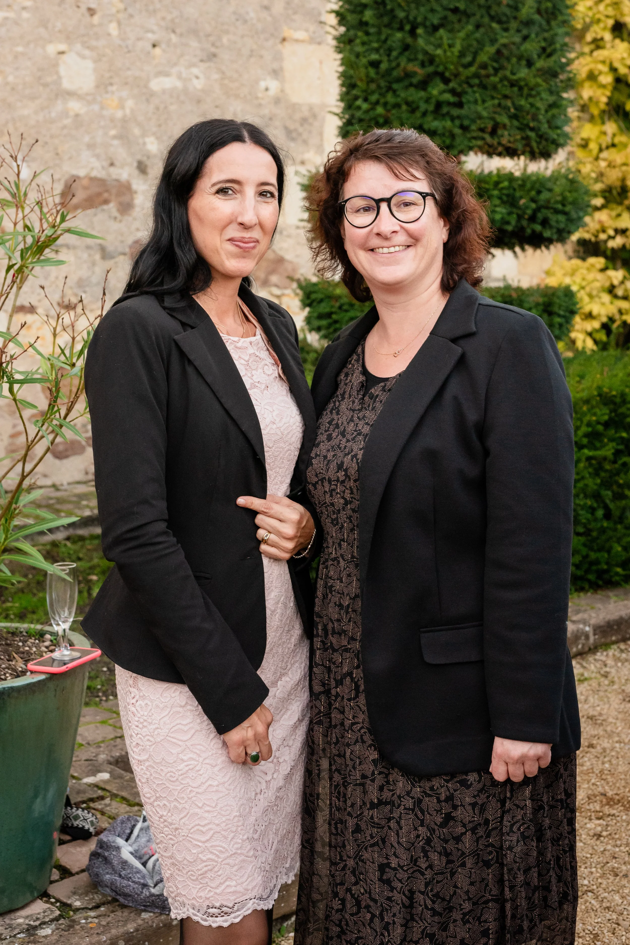 Deux femmes debout côte à côte dans un jardin, souriant vers la caméra. L'une porte une robe rose et une veste noire, l'autre porte une robe noire à motifs et une veste noire, toutes deux avec des cheveux sombres ou bruns et des lunettes. En arrière-