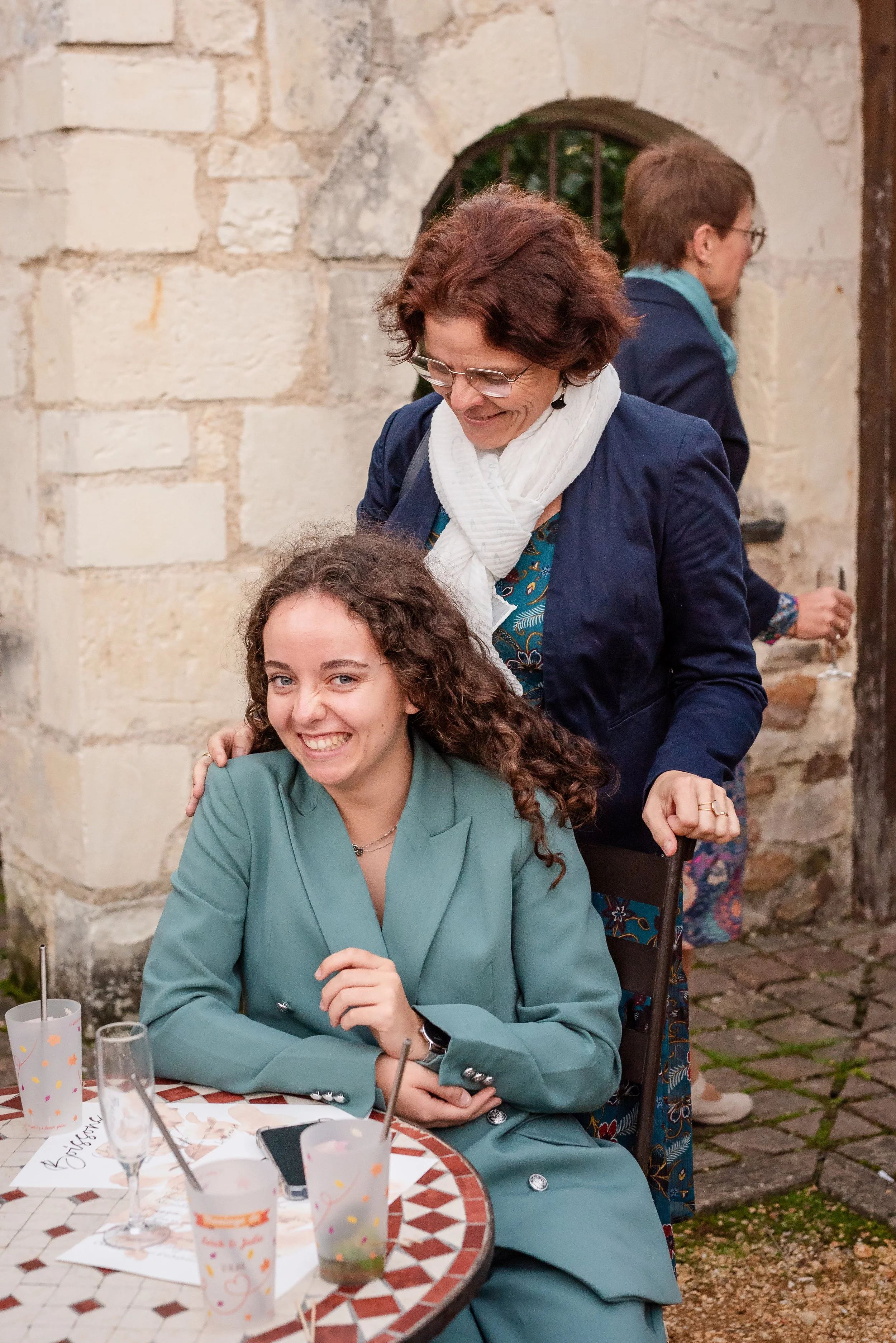 Deux femmes sont assises à une table en terrasse, en train de sourire. Une femme porte un tailleur vert et une autre un vêtement clair avec un blazer bleu. En arrière-plan, deux autres personnes discutent près d'un mur en pierre.