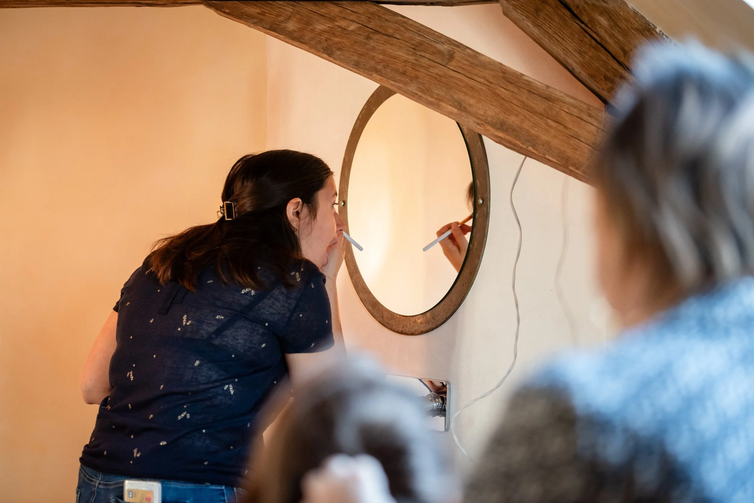 Une femme à cheveux longs, portant une chemise noire avec des motifs, se regarde dans un miroir rond fixée au mur en bois, en train de se maquiller ou de se coiffer, dans une pièce aux murs beige.