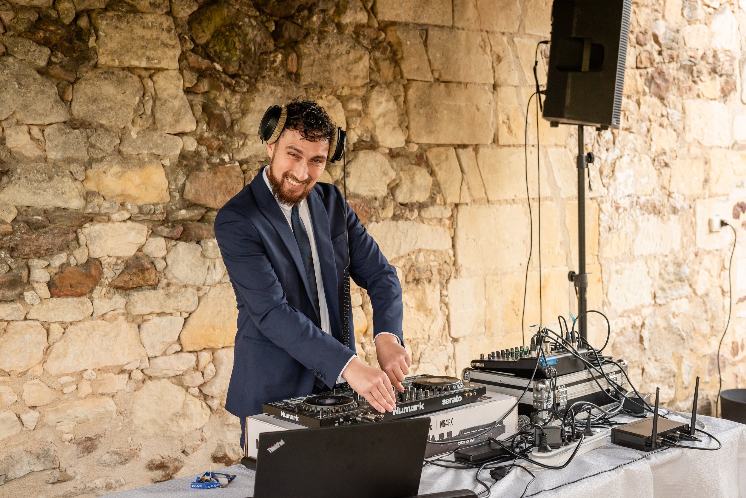 Un DJ souriant portant un costume, utilisant un lecteur de musique derrière un stand avec du matériel audio, dans une salle avec un mur en pierre visible en arrière-plan.