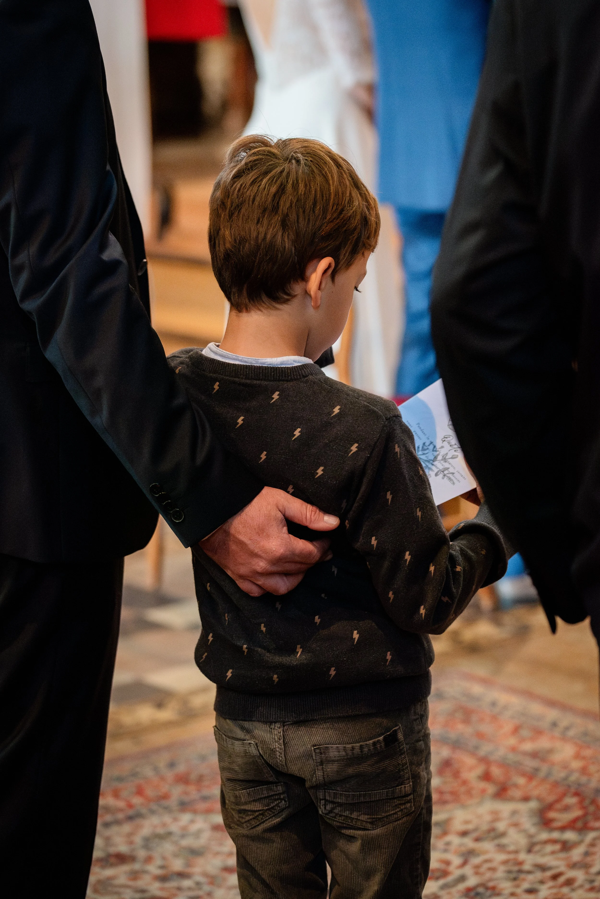 Un garçon regardant un document, entouré de deux personnes en costume, dans un lieu intérieur avec un tapis oriental.