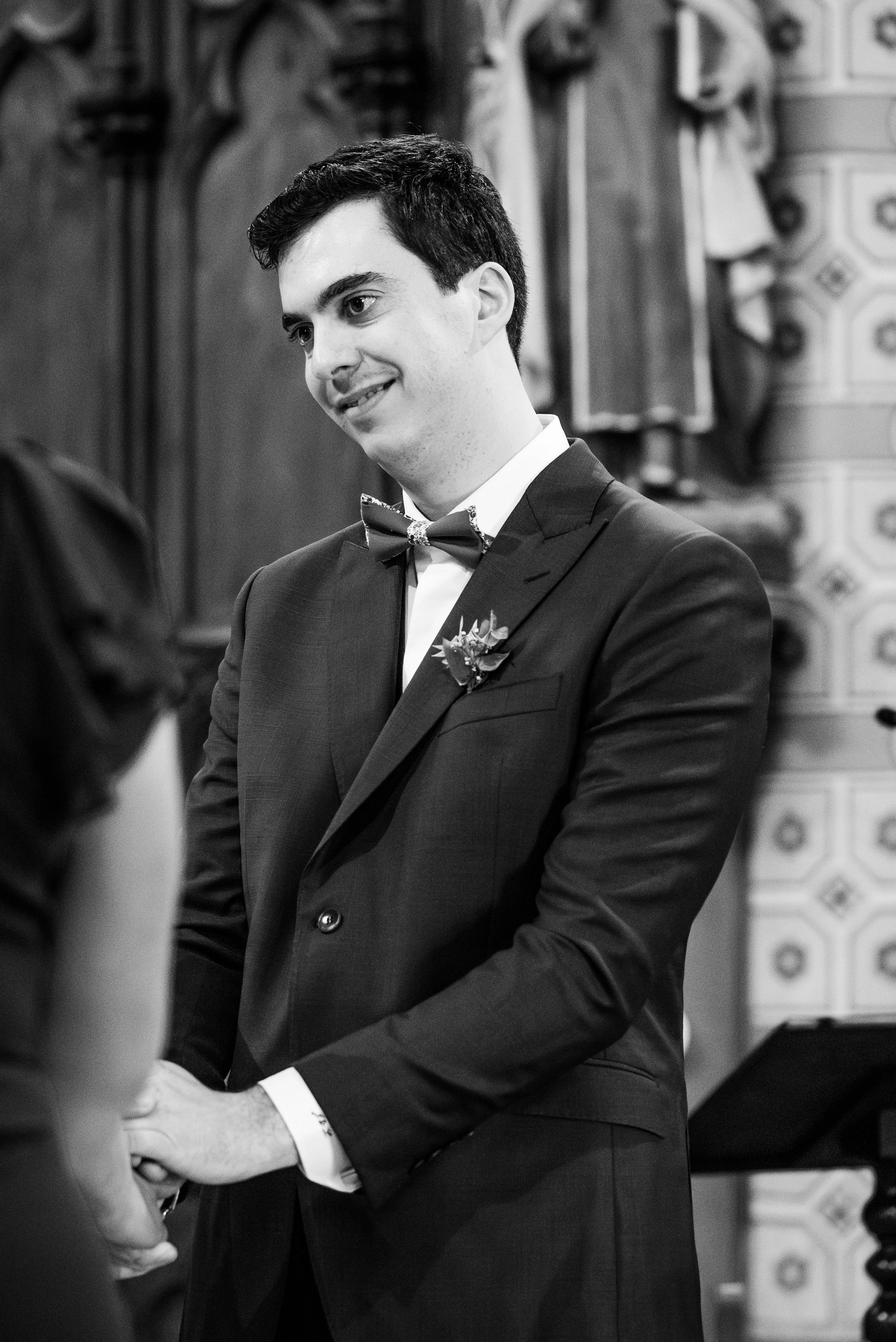 Un homme en costume noir et nœud papillon échange des vœux lors d'une cérémonie. La scène est en noir et blanc.