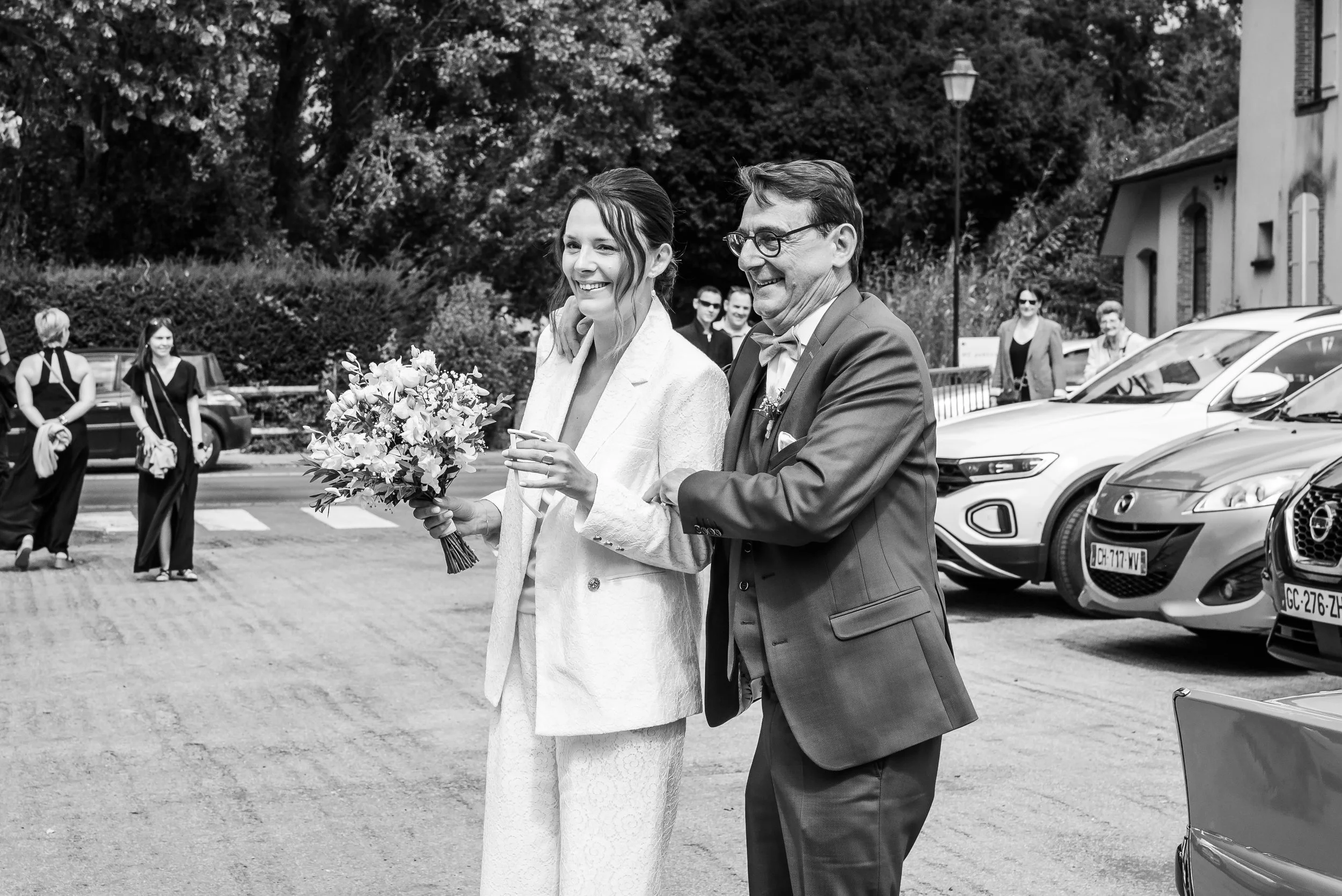 Un couple marié, la femme tenant un bouquet de fleurs, se tient devant des voitures dans une rue, entourés de personnes assistant à un mariage.