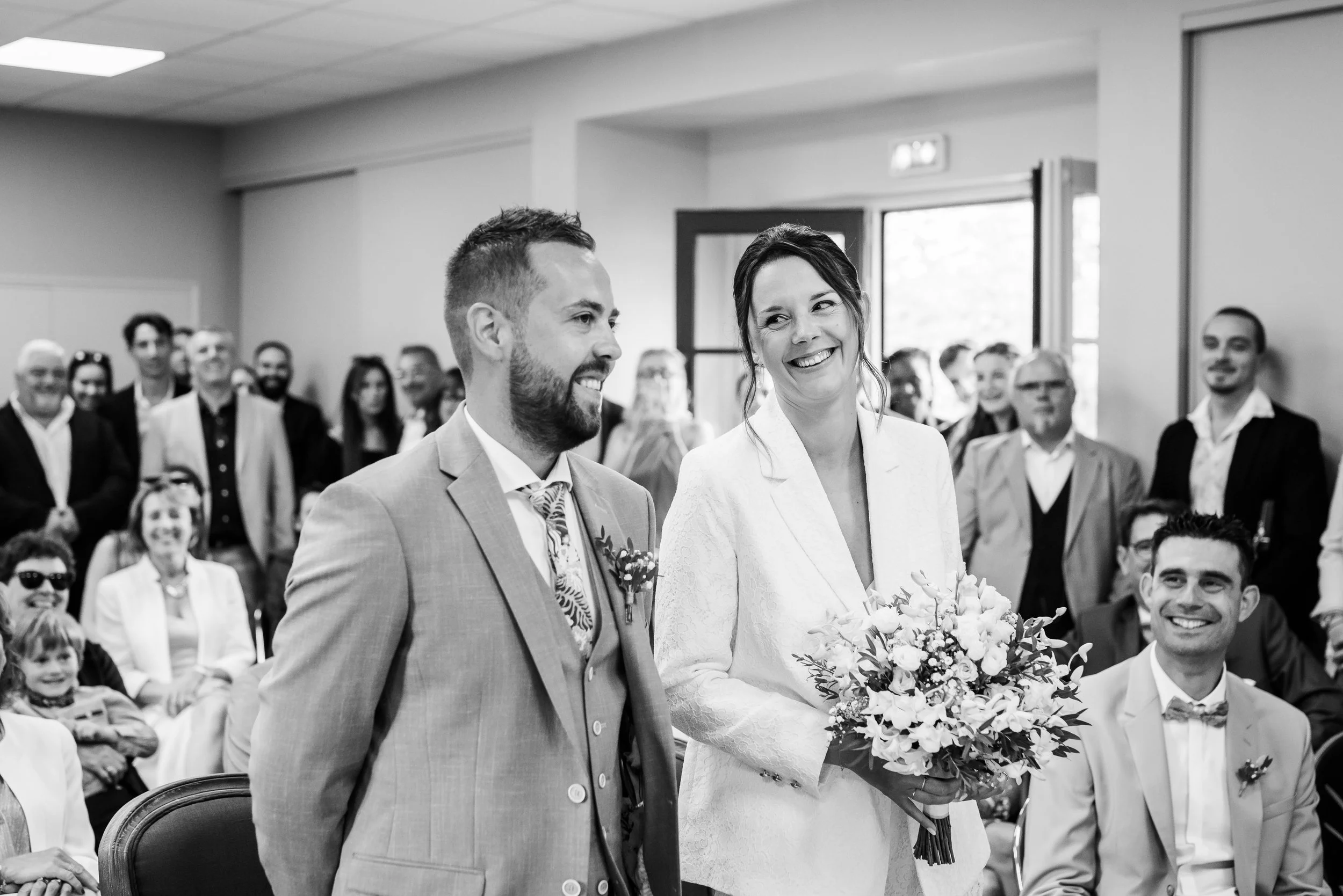 Un couple débute leur mariage lors d'une cérémonie en intérieur, entourés par des invités assis qui les regardent avec admiration, la femme tenant un bouquet de fleurs et l'homme en costume. Photo en noir et blanc.