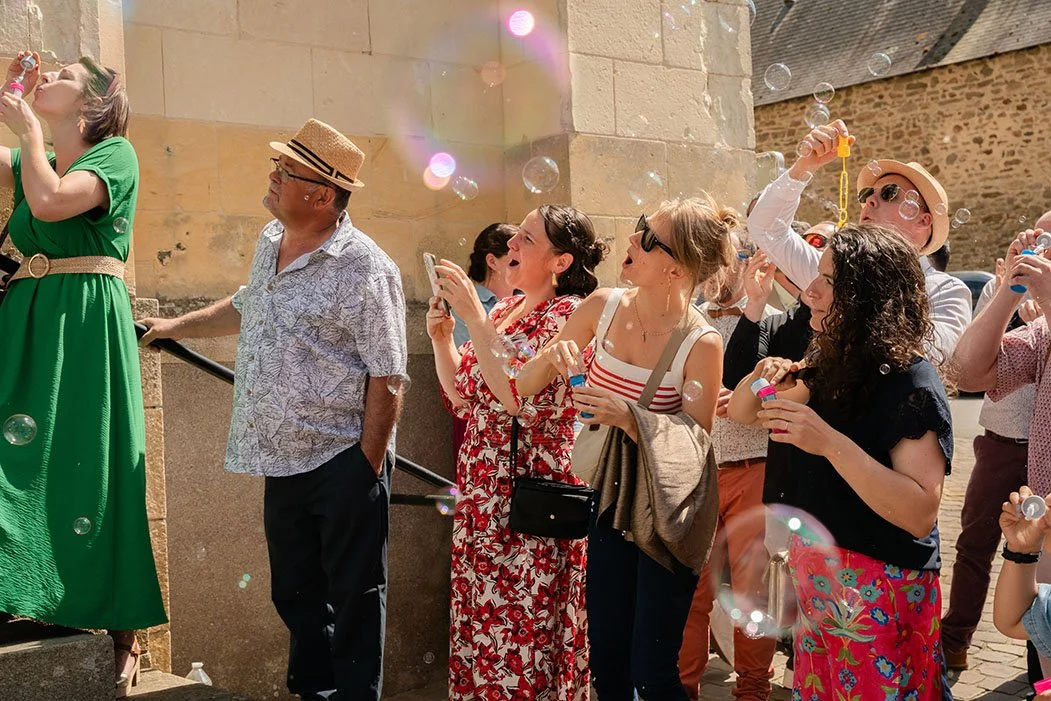 Groupe de personnes en été, regardant des bulles de savon, certains prenant des photos ou vidéos, avec un bâtiment ancien en arrière-plan.