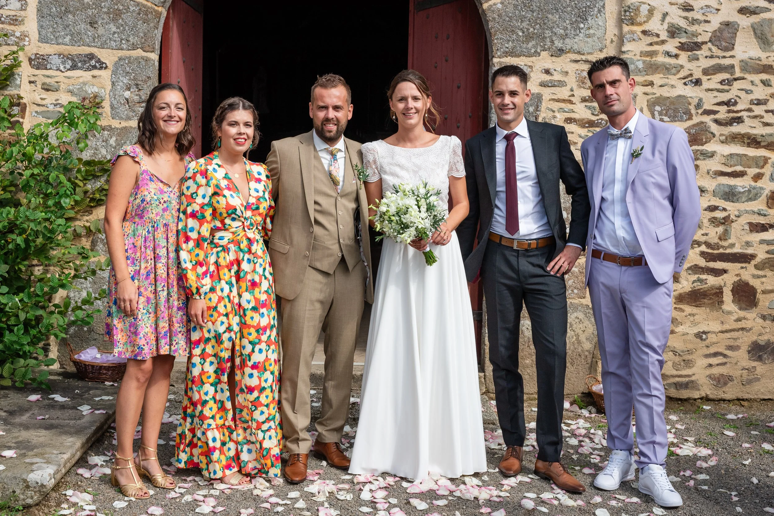 Groupe de six personnes posant devant une vieille bâtisse en pierre lors d'une cérémonie ou d'un mariage, avec des pétales de rose au sol.