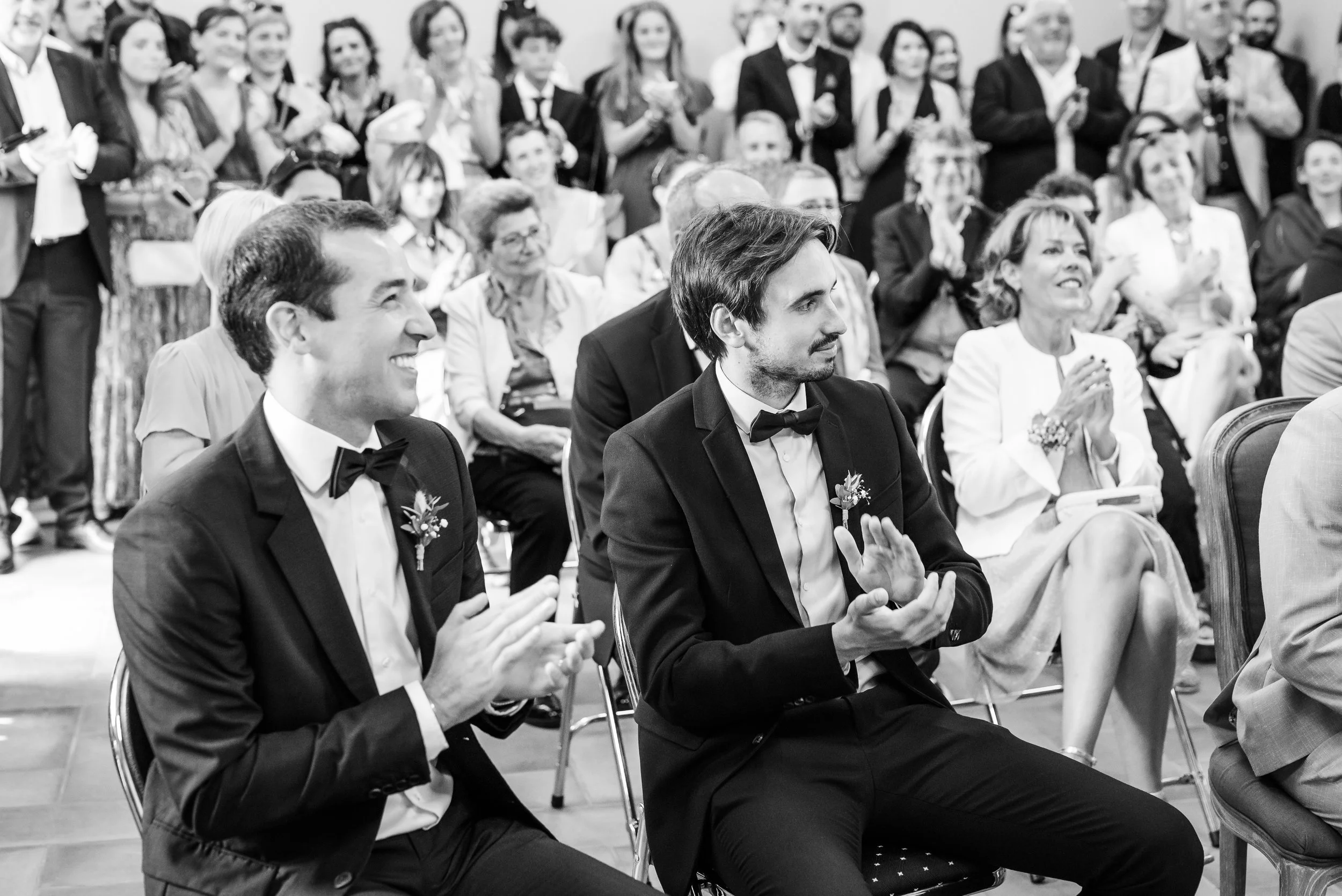 Des personnes assistent à une cérémonie, probablement un mariage, en tenant des discours et applaudissant, vêtues de costumes formels.