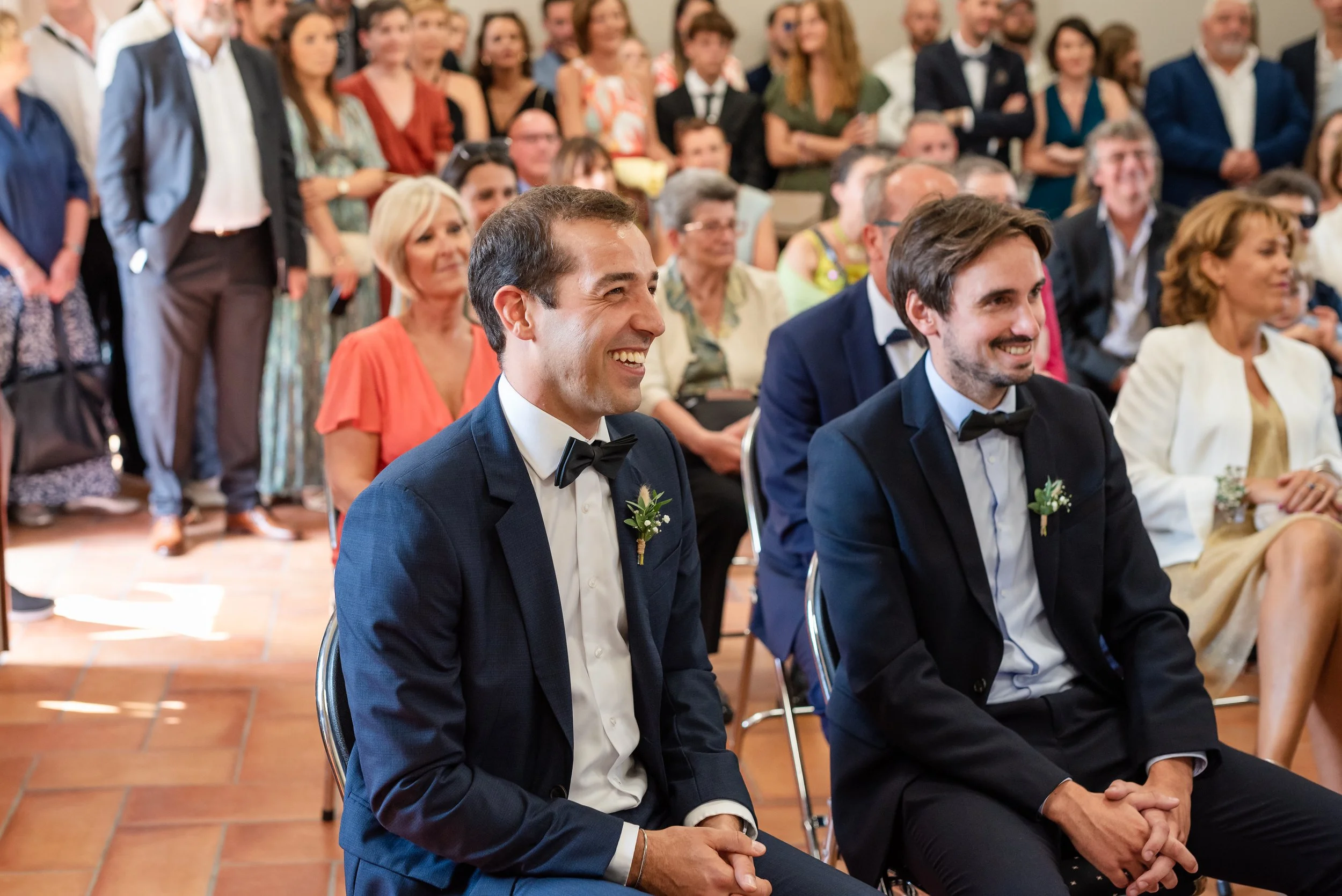 Groupe de personnes assises et debout, en gownes formelles, lors d'une cérémonie ou un événement social, avec des expressions joyeuses et souriantes.