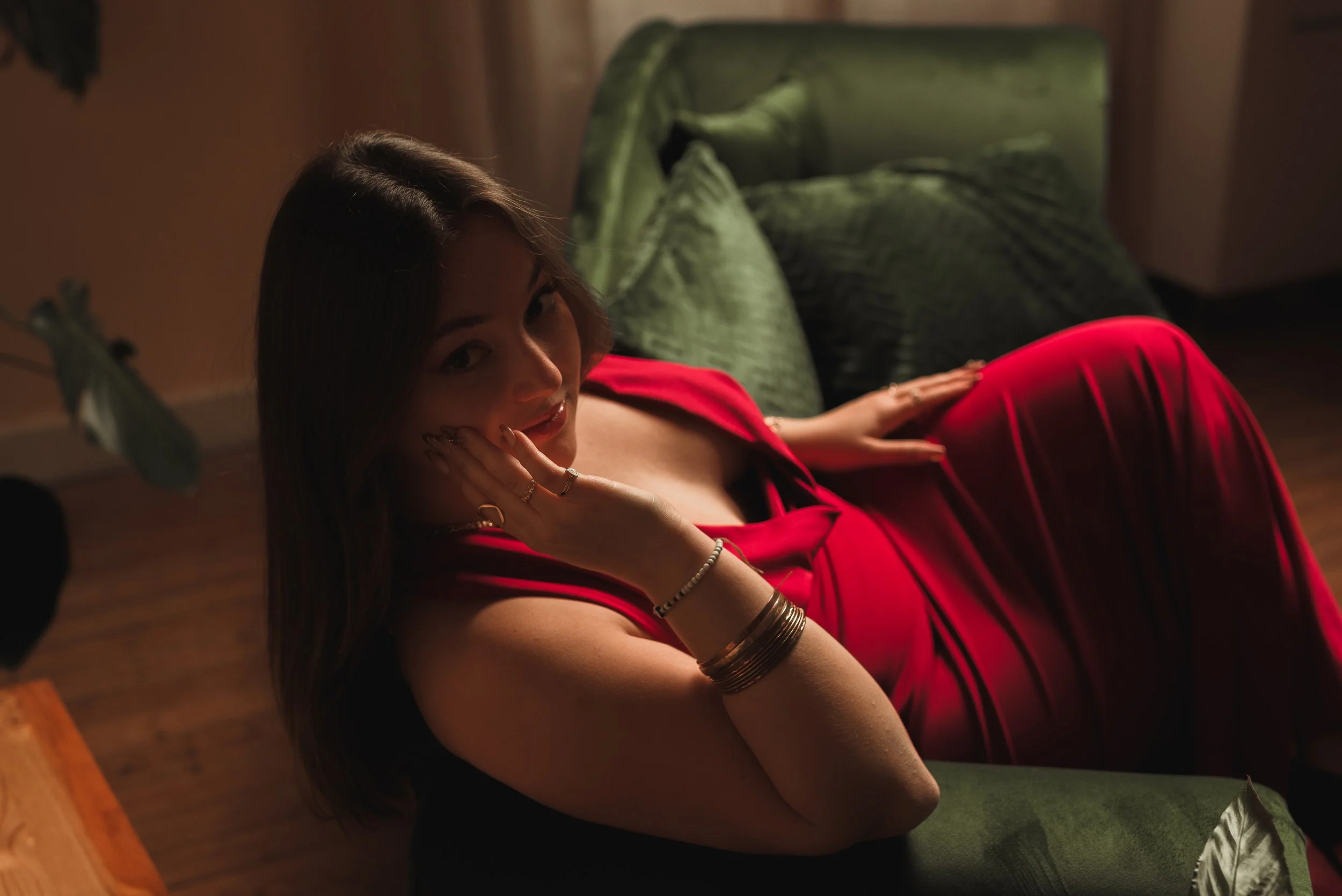 Femme avec des cheveux longs et bruns, portant une robe rouge, allongée sur un canapé vert, regardant vers l'objectif, dans une ambiance tamisée.