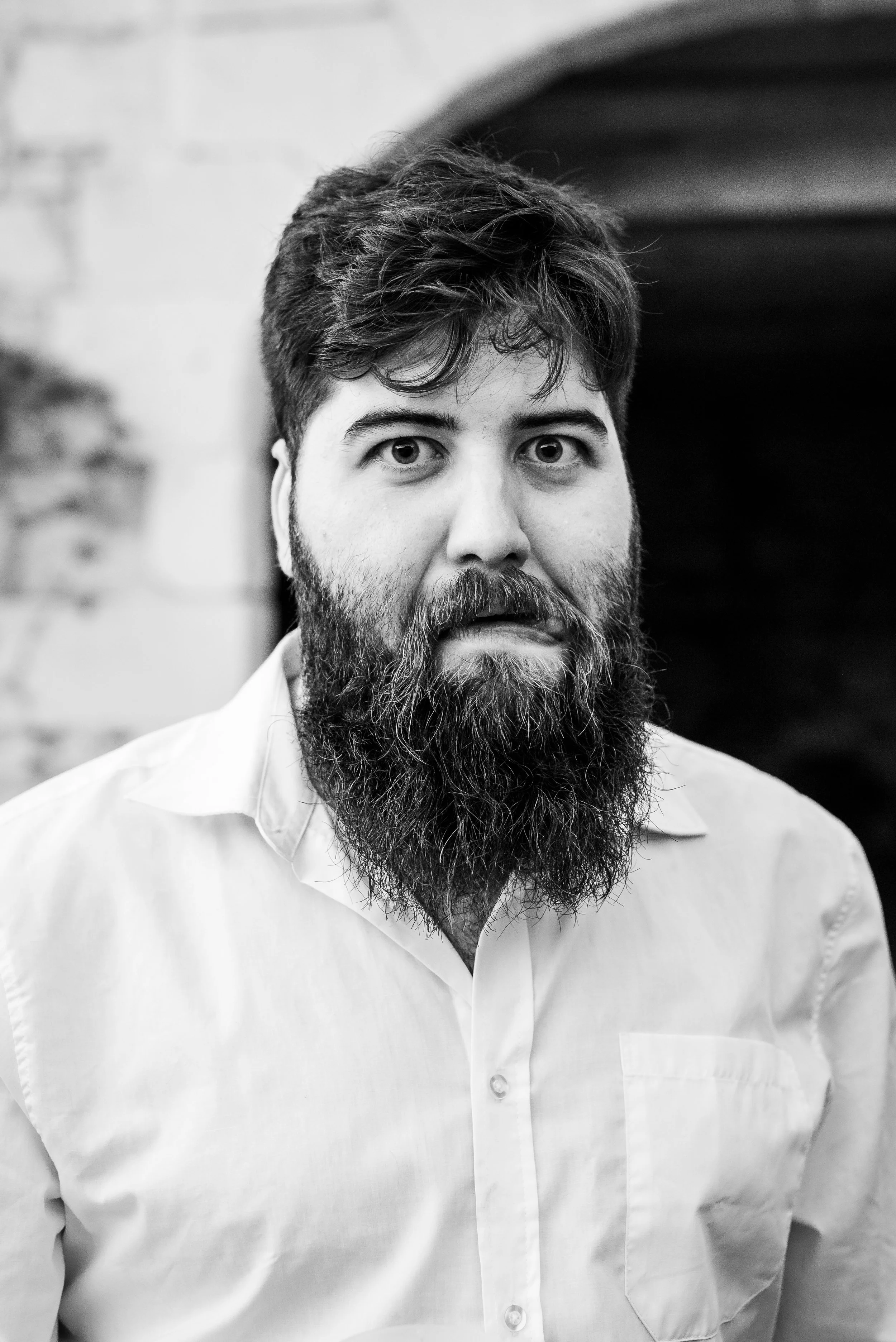Homme avec une barbe épaisse, cheveux courts, portant une chemise blanche, regardant devant lui, en noir et blanc.
