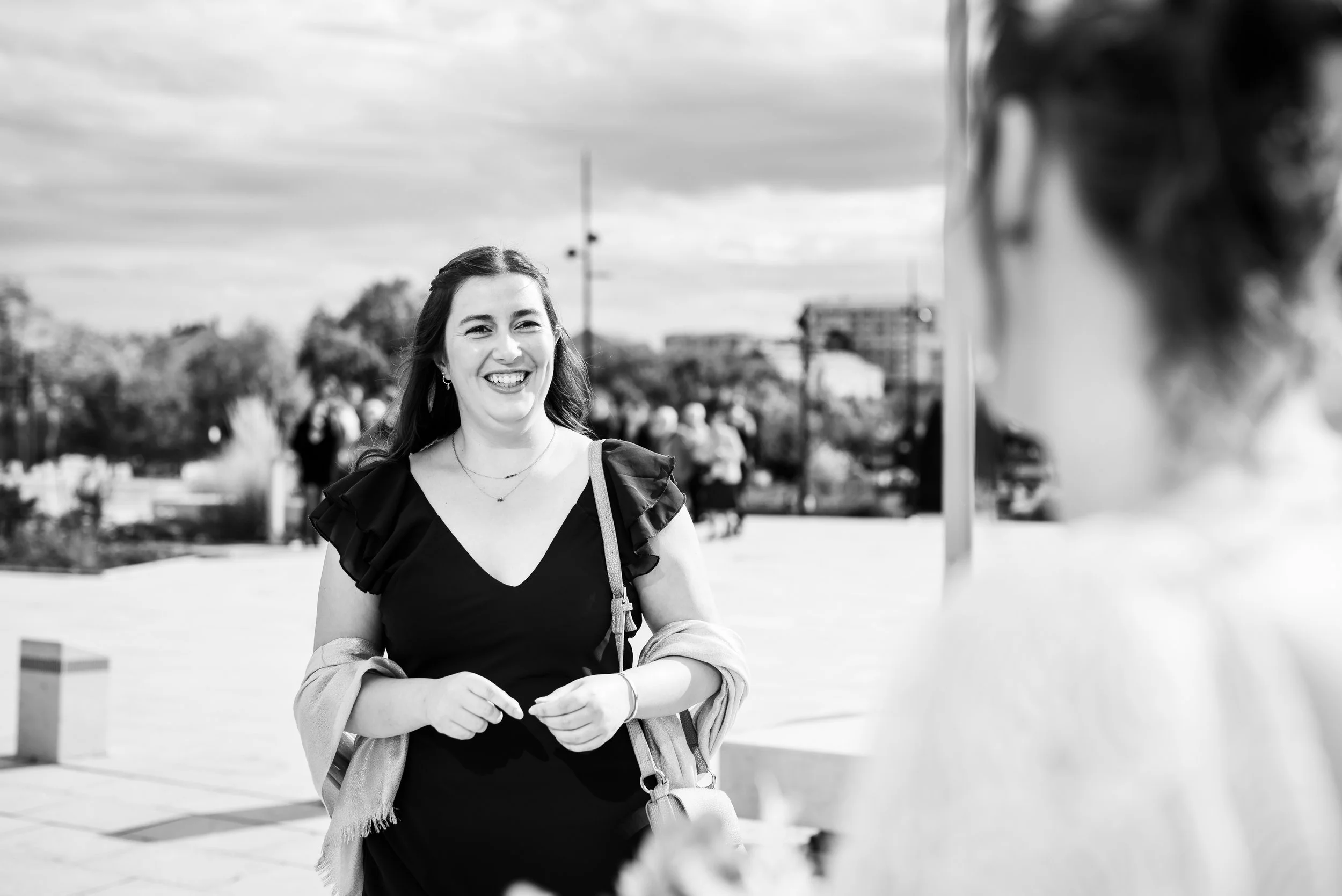 Une femme souriante en vêtements noirs, portant un sac, avec d'autres personnes en arrière-plan dans un espace extérieur, en noir et blanc.