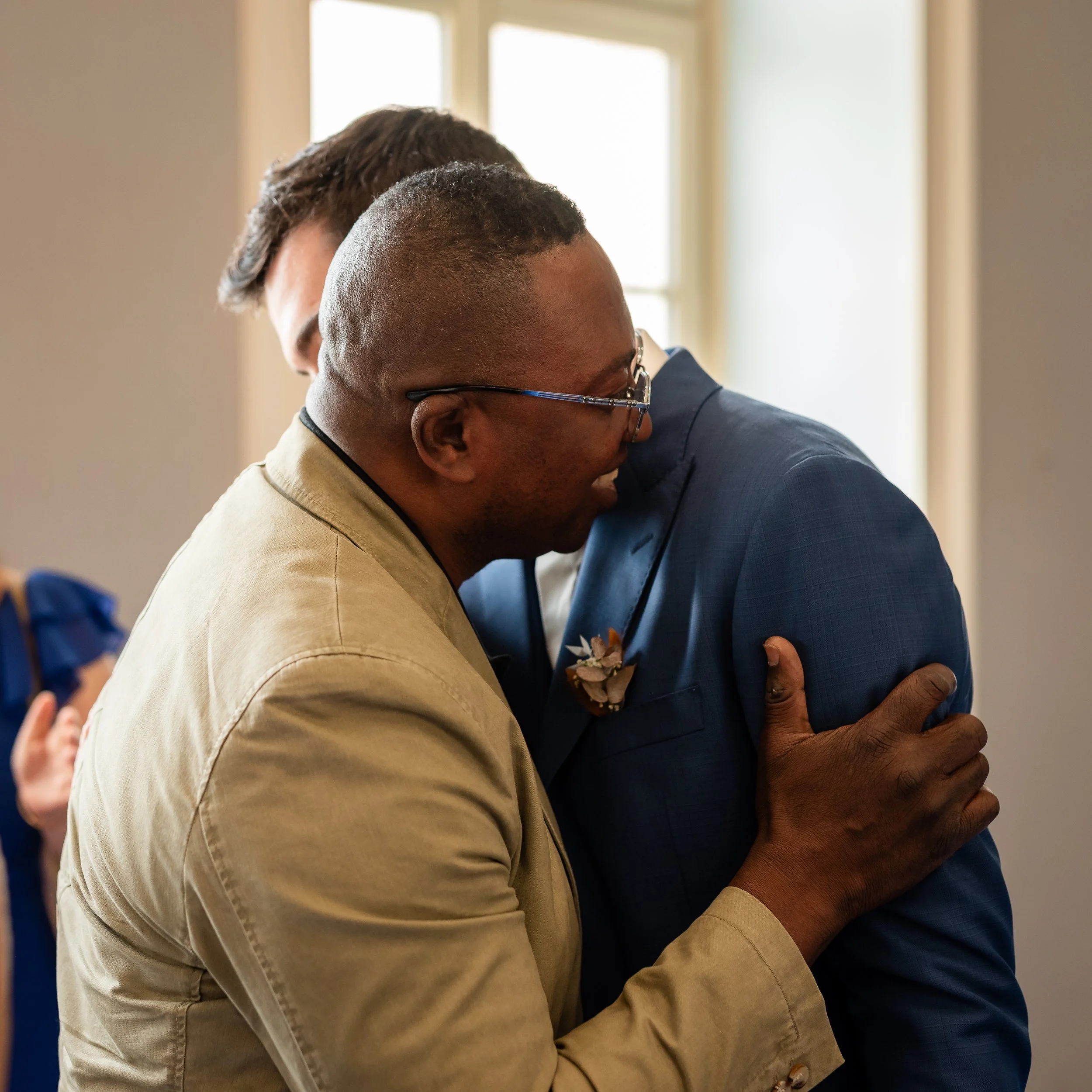 Deux hommes s'embrassent dans une accolade chaleureuse, l'un portant des lunettes et l'autre habillé en costume bleu, dans une pièce baignée de lumière naturelle.