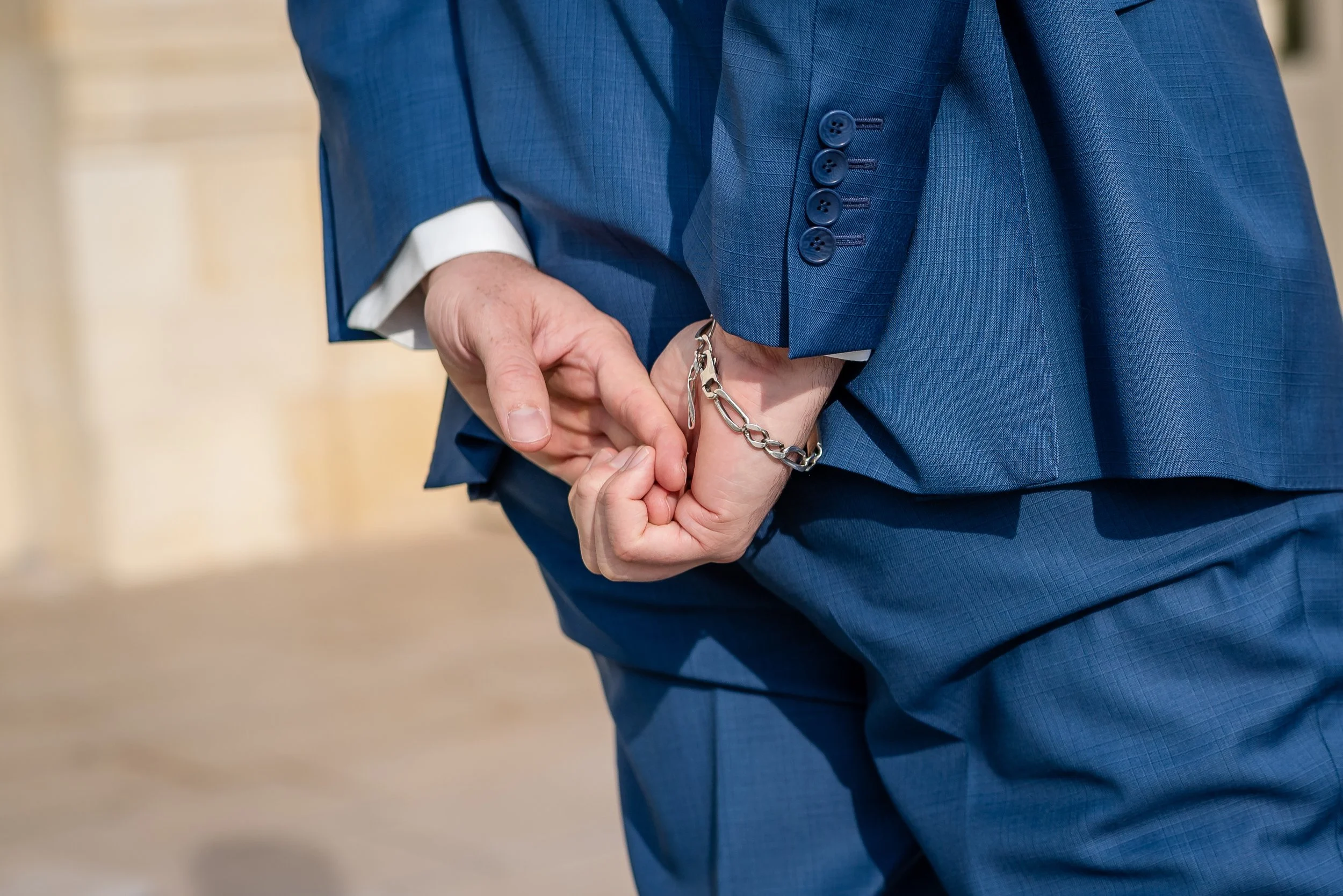 Une personne en costume bleu tient ses mains derrière le dos, porte une montre ou bracelet en chaîne argentée.