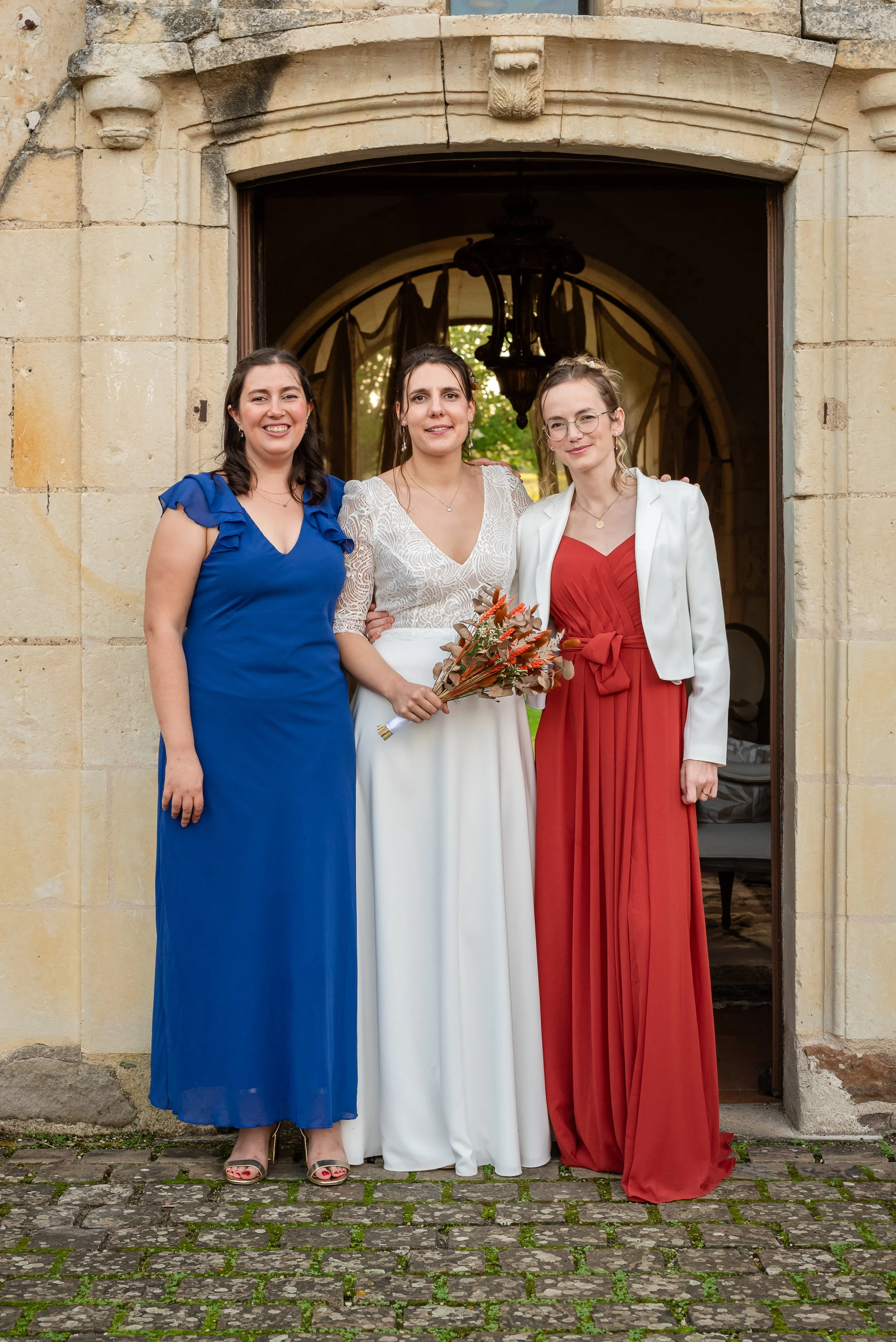 Trois femmes posant devant une porte en pierre, une extérieur. La femme au centre porte une robe blanche avec un bouquet de fleurs, probablement une mariée. Les autres femmes portent des robes longues, une en bleu et l'autre en rouge, semblant être d